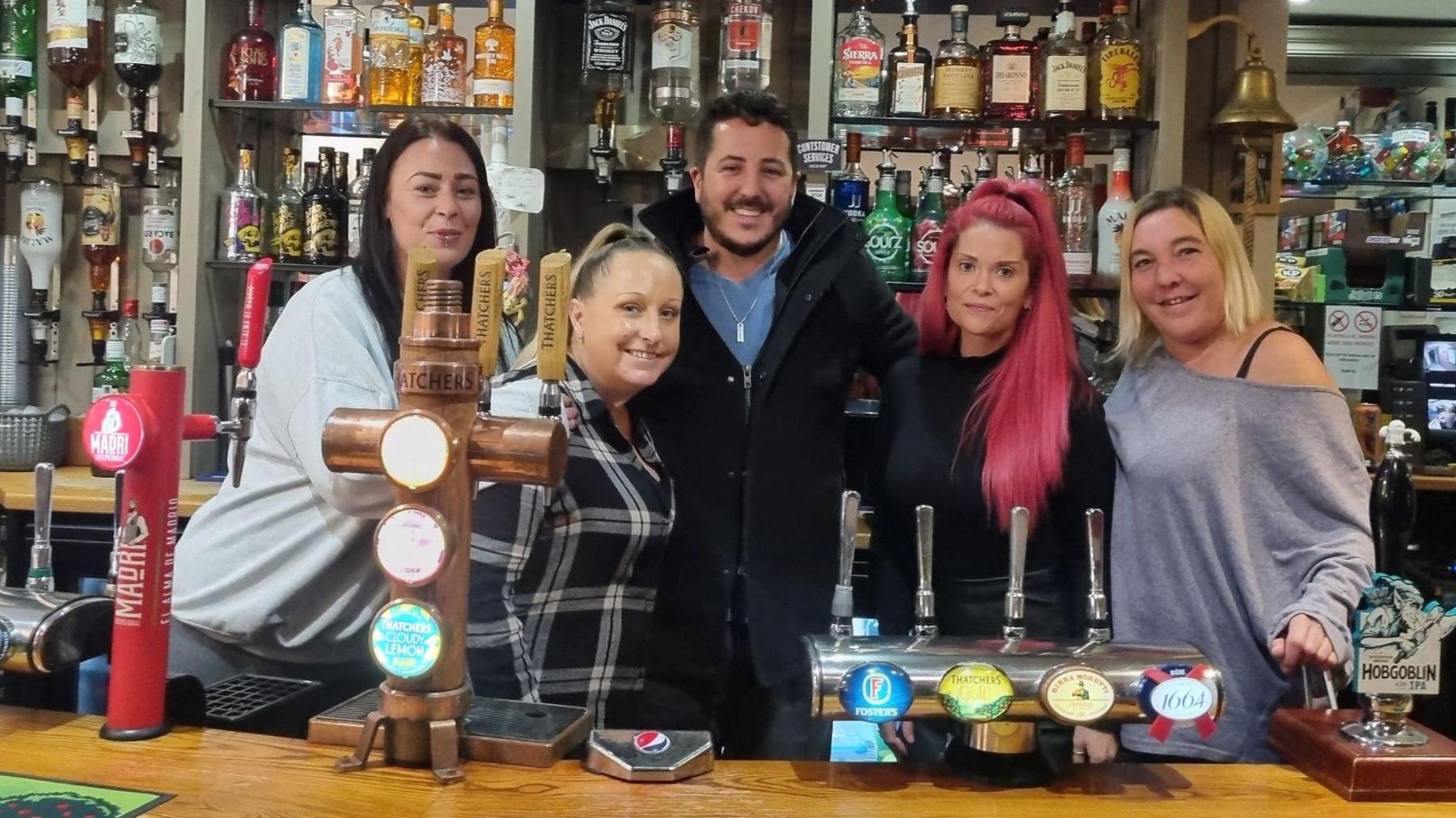 A pub bar with bottles bahind and the team. Dale is in the centre with two women either side (four in total) making up the pubs staff.