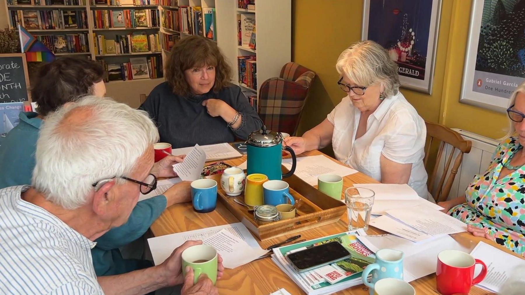 Ginny Sealey with members of a book club sat around a table. Ginny is at the head of the table in a blue jumper. There are two women and two men sat with her. There are pieces of paper and cups of tea on the table.