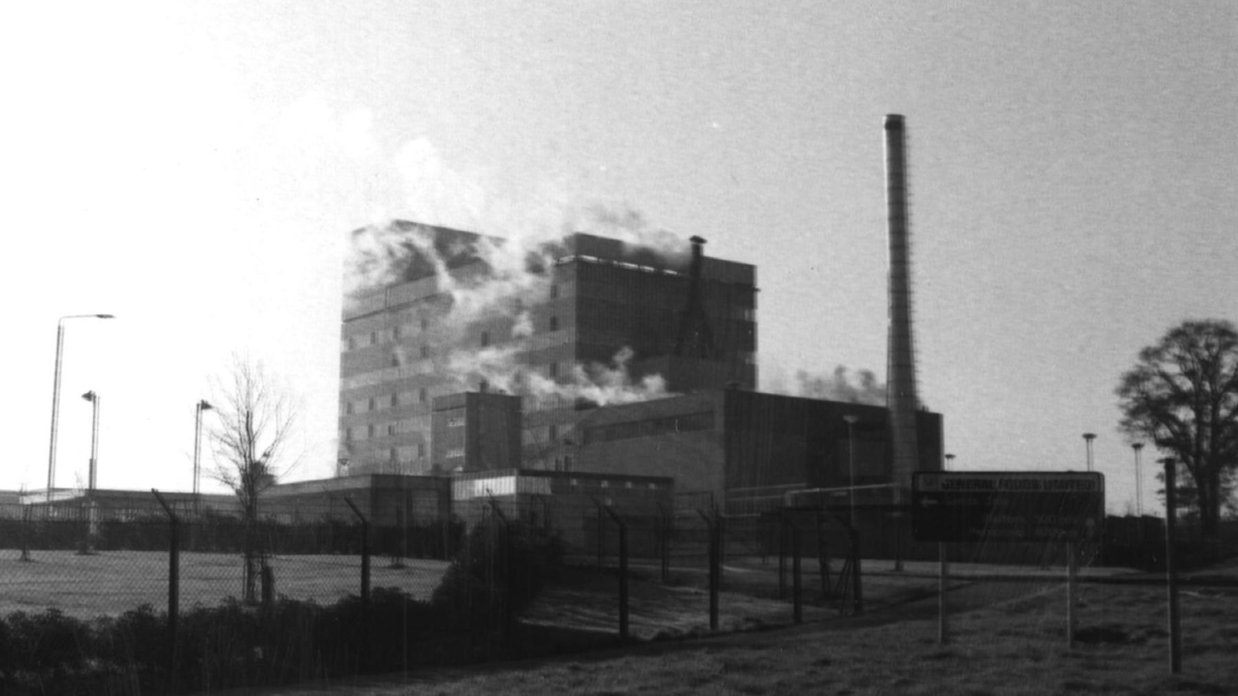 A black and white image of the plant and a chimney, among fields.