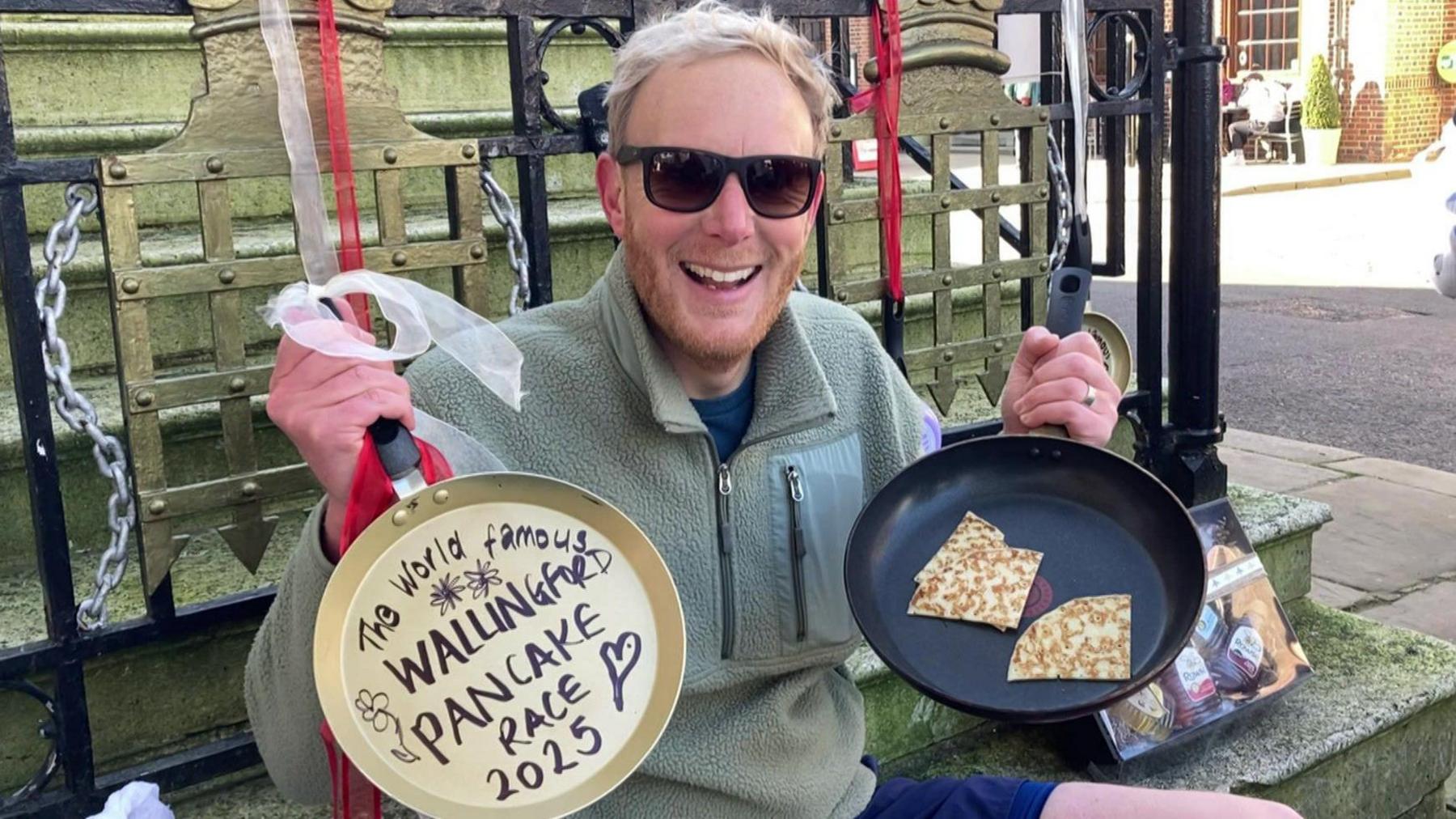 Ben Hodgkinson smiles at the camera. He is sitting on a set of steps in Wallingford town centre. He has short blonde hair and a short ginger bear, wears black Ray Bans with a pale green zipped up fleece and blue shorts. He is holding a frying pan in his left hand with two triangular folded pancakes inside and in his right hand he's holding a golden frying pan, which has a red and white ribbon tied to the handle and the pan has writing which says, "The world famous Willingford pancake race 2025"