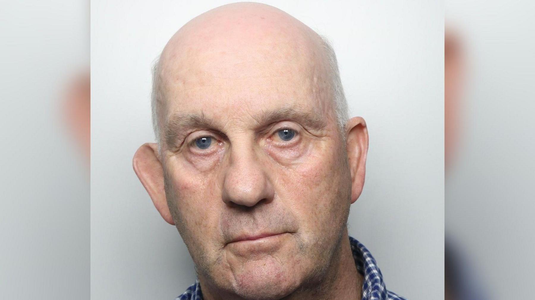 A police custody picture showing a close-up image of a man with short grey hair on the sides and a bald head. He is wearing a blue and white checked top. 