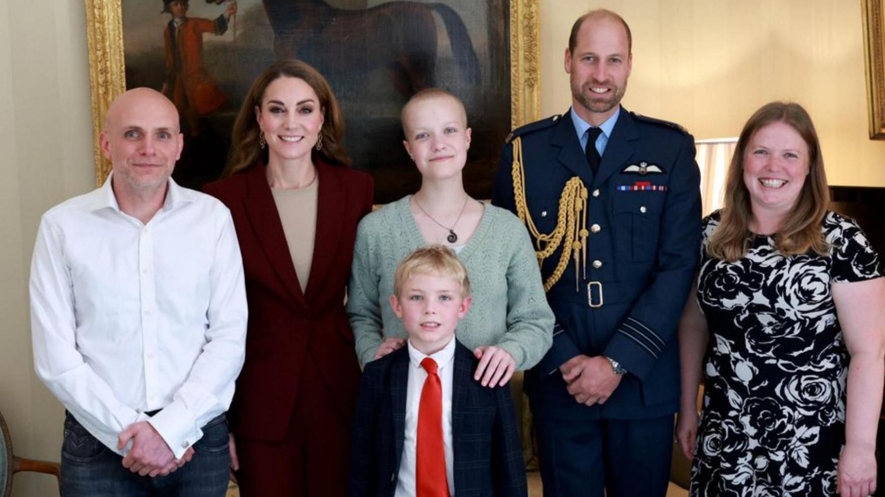 Liz Hatton, pictured with the Prince and Princess of Wales. The Princess, who is smiling at the camera, wears a burgundy blazer and beige top. She stands next to Ms Hatton, who is wearing a light green jumper and necklace. Her hands rest on the shoulders of a young, blond boy in front of her, who is wearing a dark suit and red tie.