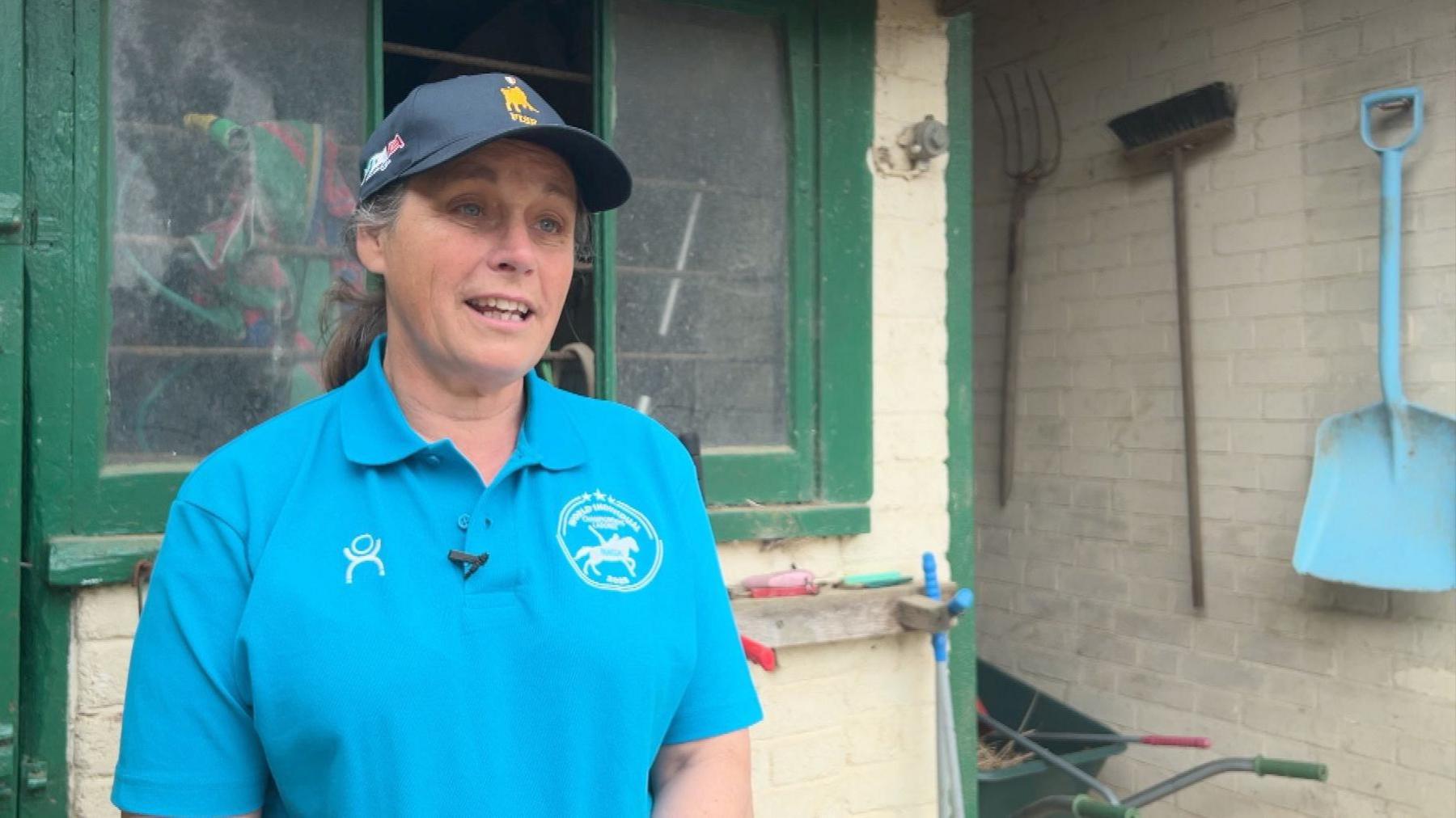 There is a woman on the left-hand side of the image, wearing a bright blue collared top with a white logo on either side of her chest. She is also wearing a dark blue cap with a yellow logo on it. Her grey hair is packed backwards in a ponytail and she is angled towards the right-hand side of the image. There is a building behind her, with green painted around the window frame and garden tools can be seen on the right side of the image, including a light blue shovel, a broom and a pitchfork.