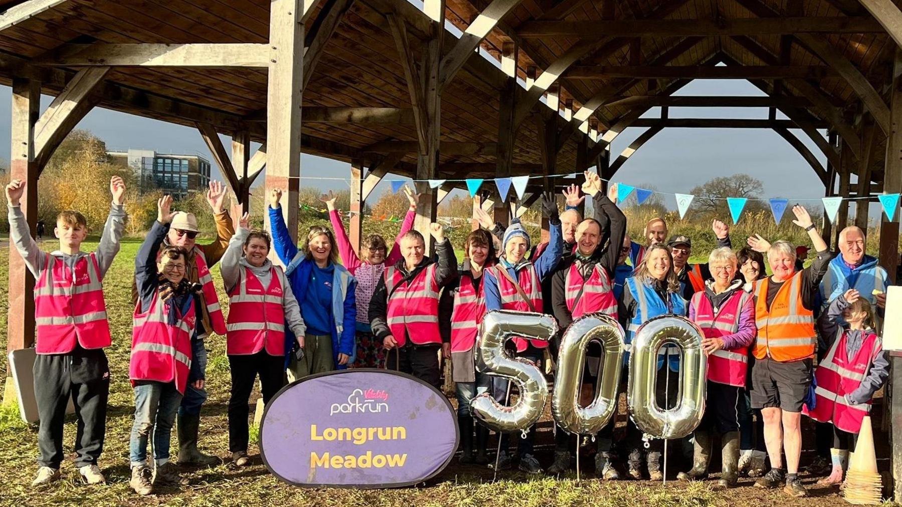 A row of 19 people, mostly in pink hi-vis jackets have their hands in the air and are smiling at the camera. They're stood underneath a barn-like structure, and in front of them is a blue sign which says 'Longrun Meadow'.