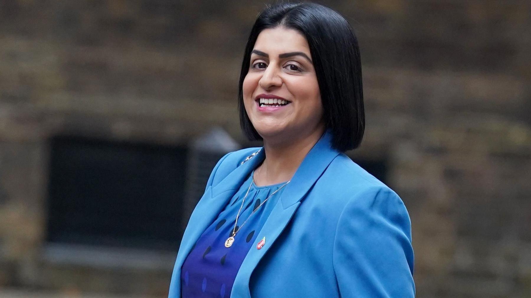 Justice Secretary Shabana Mahmood wears a turquoise blazer and indigo and turquoise dress, smilling at the camera side-on.
