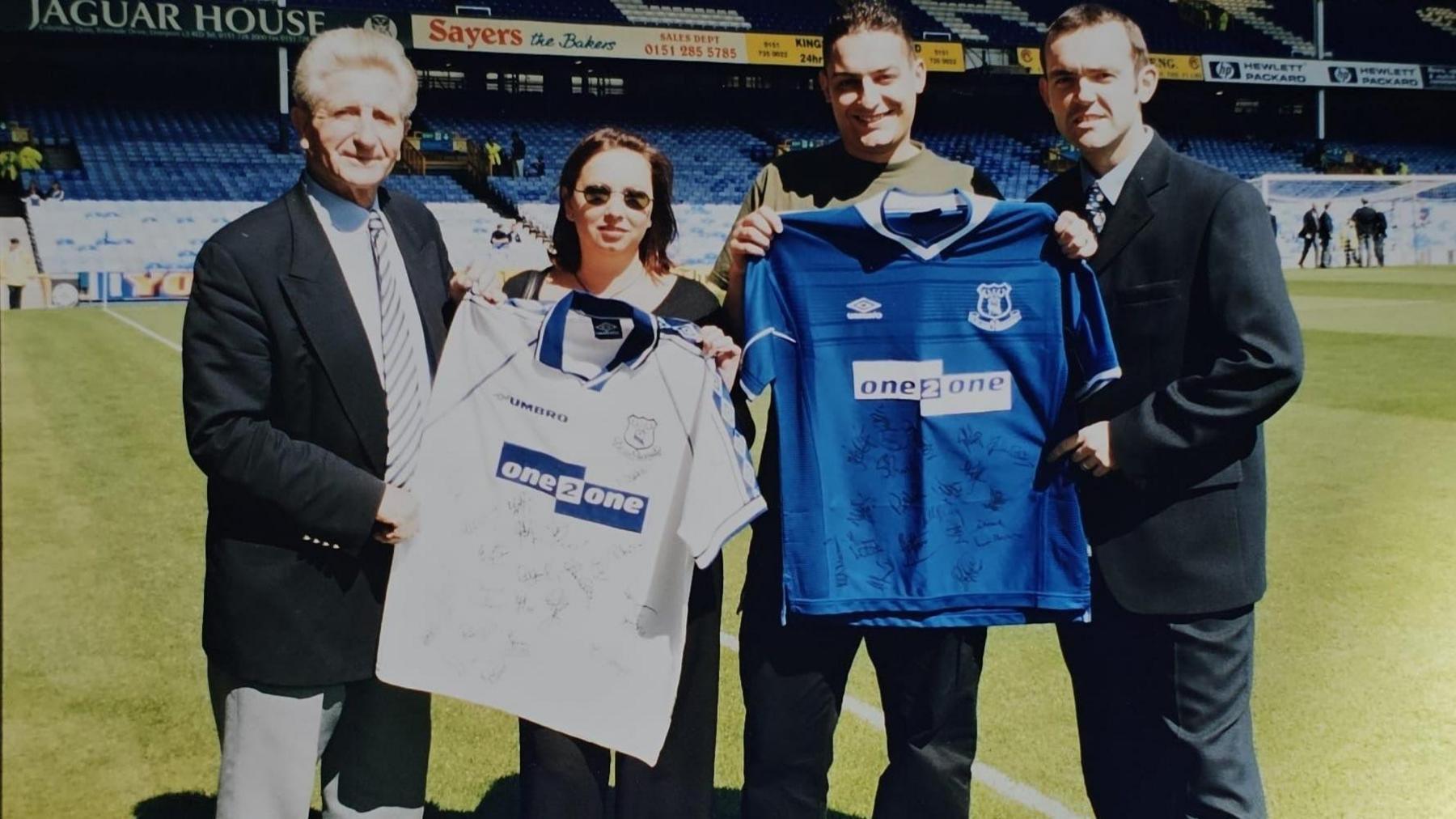 Del and Aly Thornton are standing next to each other holding two signed Everton shirts - one blue and one white. They are situated pitch side with two Everton officials standing either side of them.