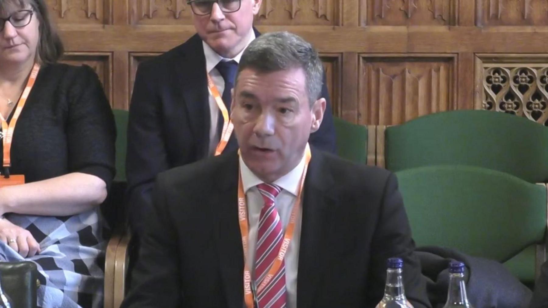 Lawrence Gosden is wearing a black blazer, white shirt and pink tie, and is sat down answering a question.