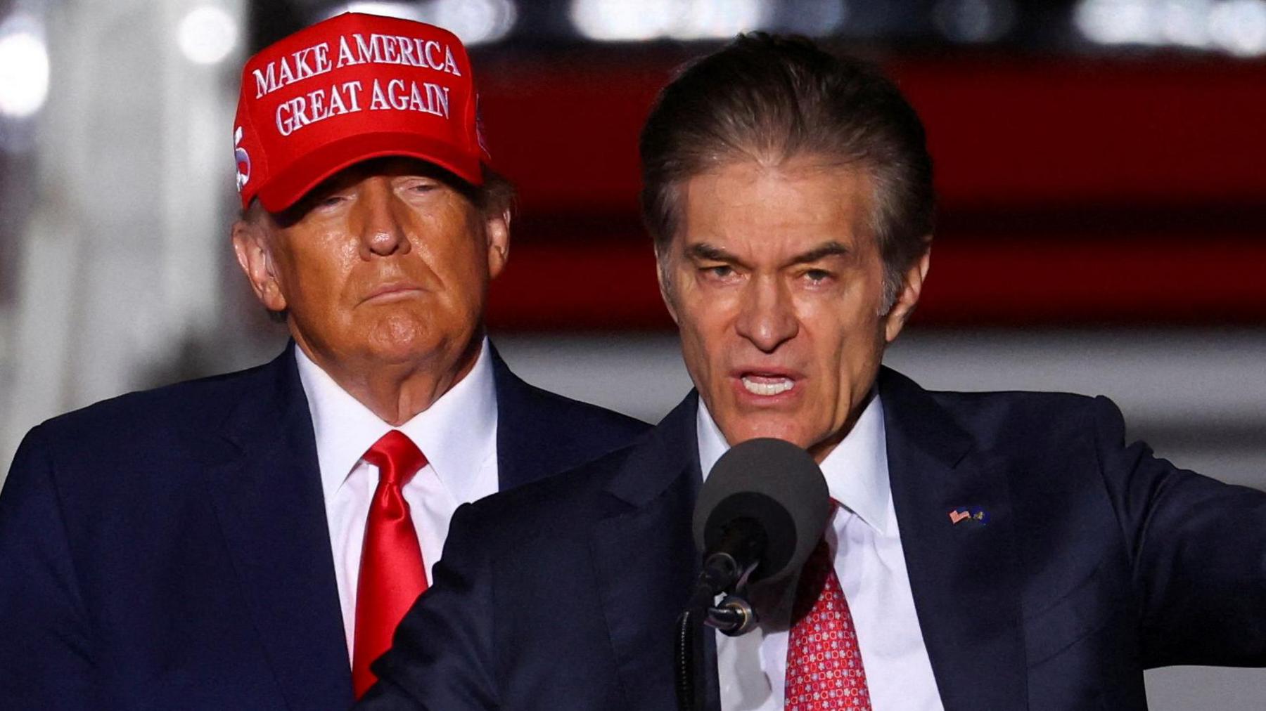 Mehmet Oz speaking into a microphone and gesturing as Donald Trump behind him looks on, wearing a red "Make America Great Again" hat. 