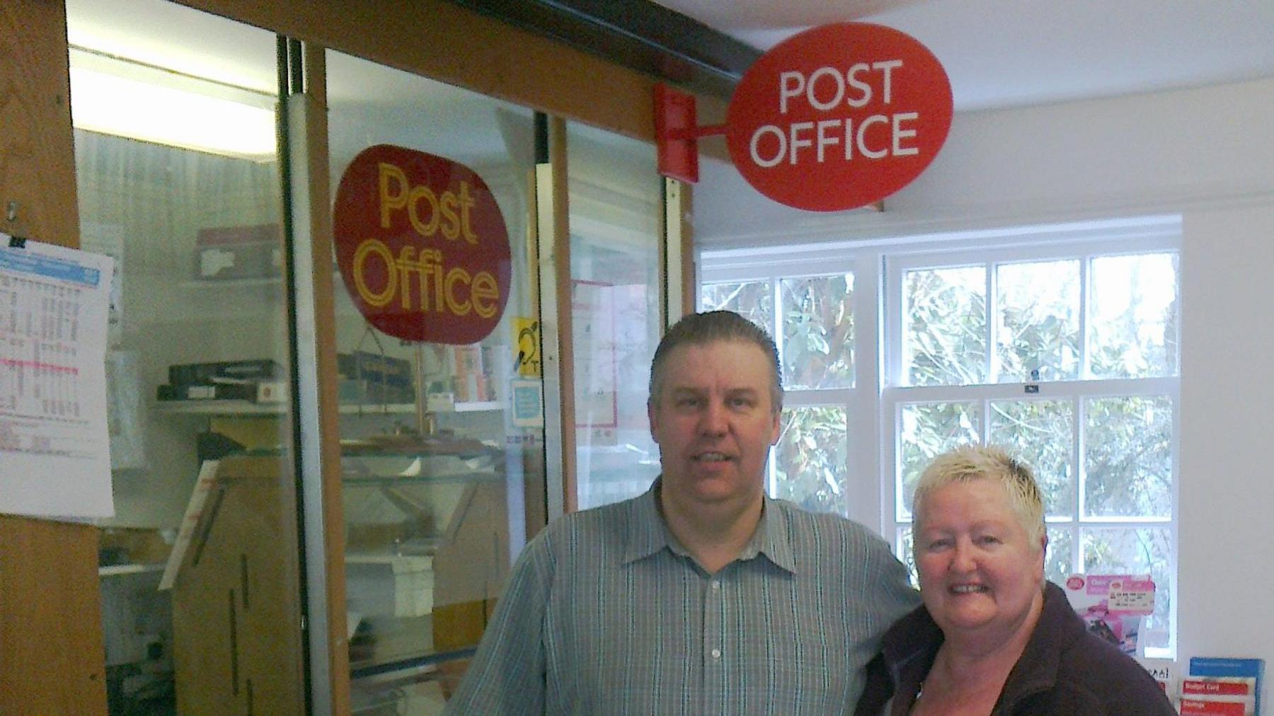 A man wearing a shirt standing next to a woman with short blonde hair. They are standing in front of a Post Office kiosk.