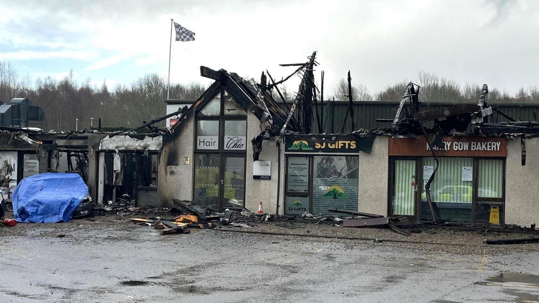 The shops have been gutted by fire. Wooden rafters and gable ends are charred. The car involved in the incident is under a blue waterproof cover.