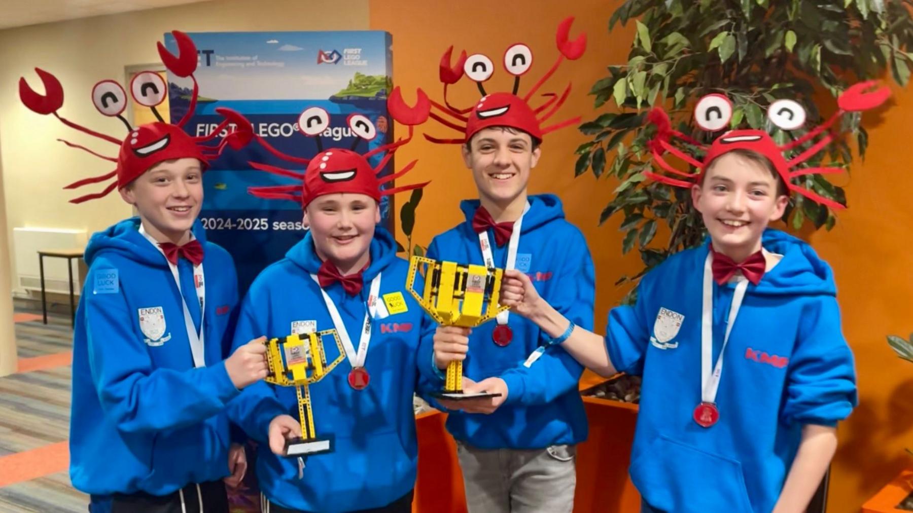 Four boys in matching blue hoodies and red hats in the shape of crabs hold two yellow trophies made from Lego bricks. They stand in front of an orange wall and large indoor plants.