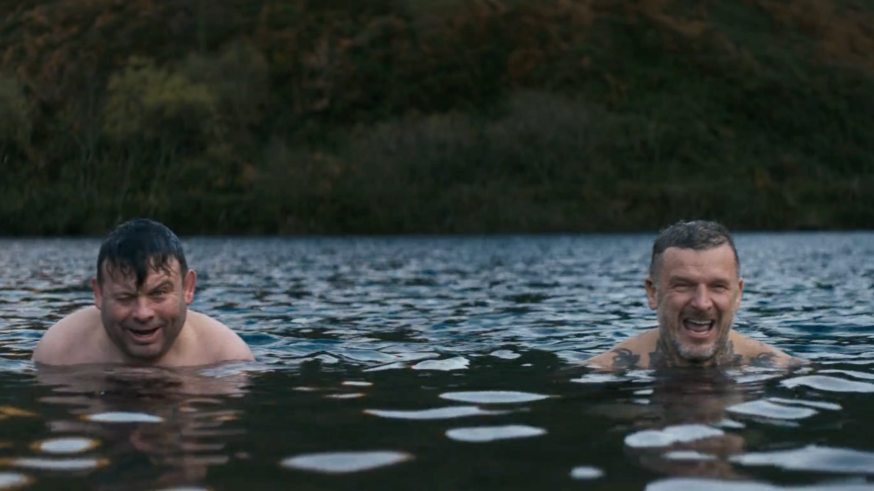 Mathew Pritchard (right) photographed with Dirty Sanchez co-star Mike "Pancho" Locke. They both sit in Brombil Reservoir, Port Talbot. Pritchard has grey short hair, a grey stubbled beard and smiles at the camera. Pancho has black hair which is wet slicked downwards onto his forehead. He holds one eye shut and smiles. A forest of pine trees can be seen behind the reservoir in the background.