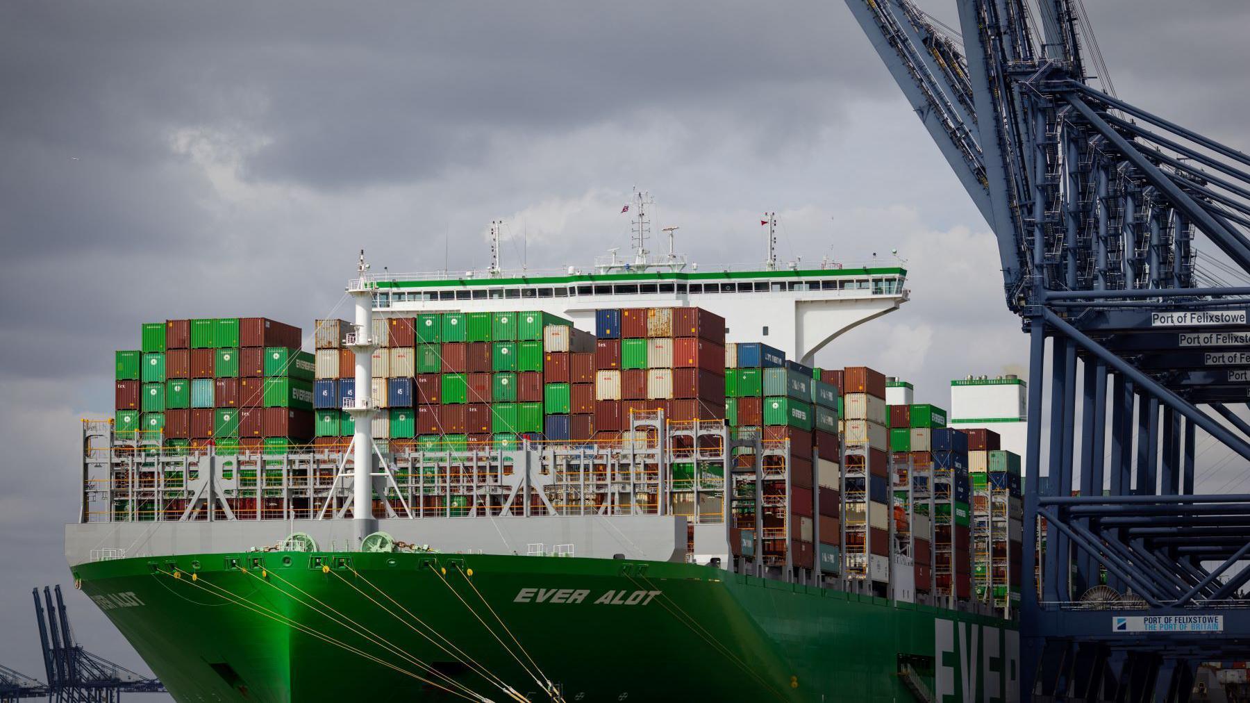 A large green shipping boat with hundreds of containers on it, which are mostly brown and green. There is a huge blue crane to the right of the frame. It is cloudy above.