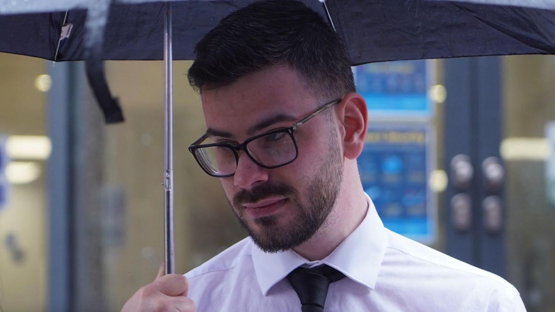 A man with dark hair and a beard, wearing black-rimmed glasses, wearing a white shirt and black tie, holding a black umbrella.