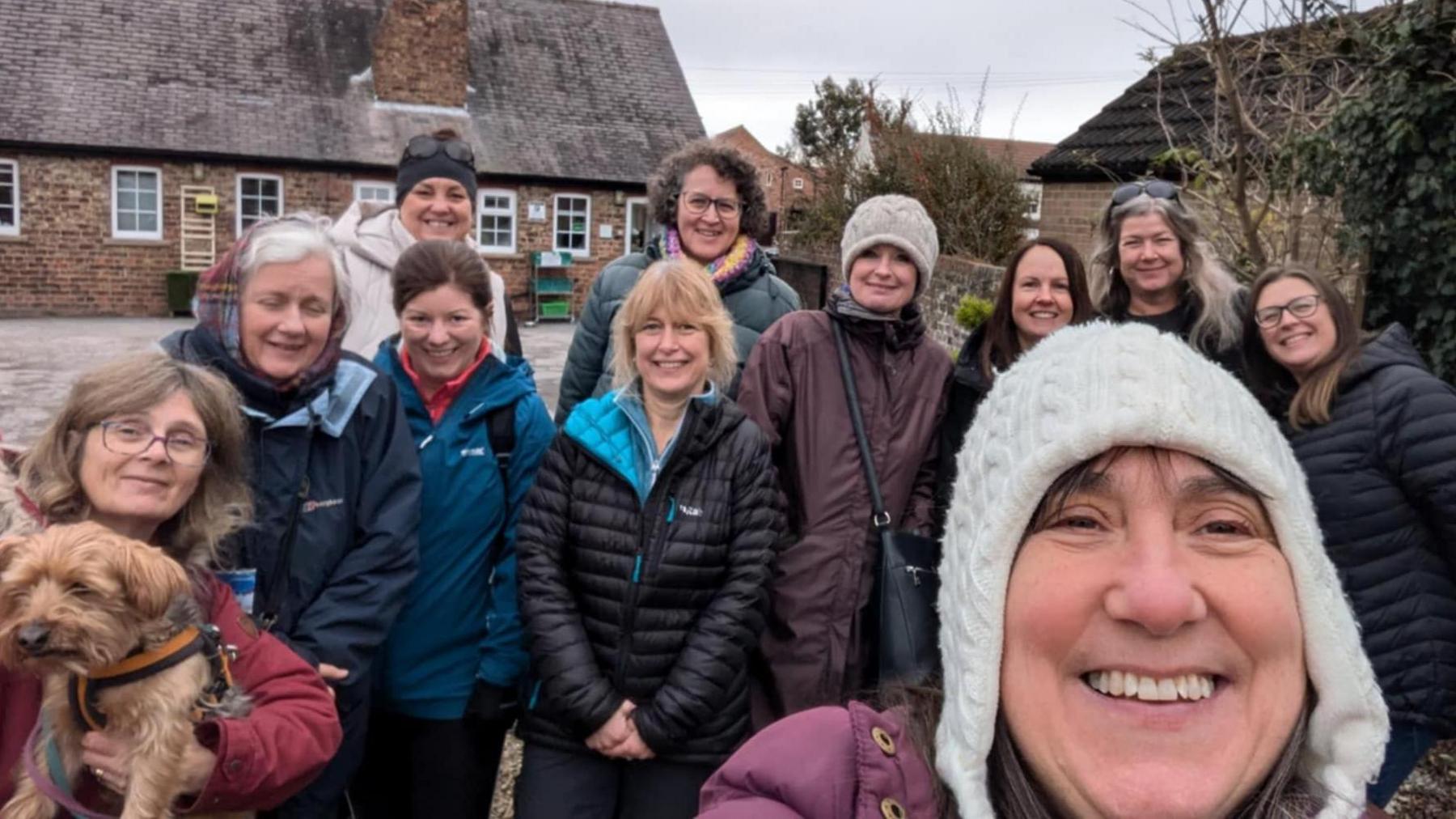 A group photo of 11 women in coats and a dog on a walk. Some are also wearing woolly hats.