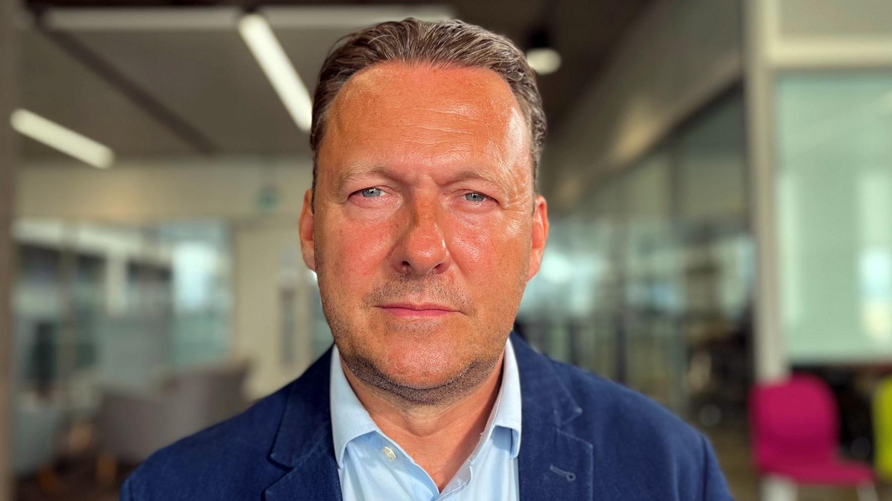 A middle aged man in a light blue open necked shirt an darker blue jacket stares directly at the camera