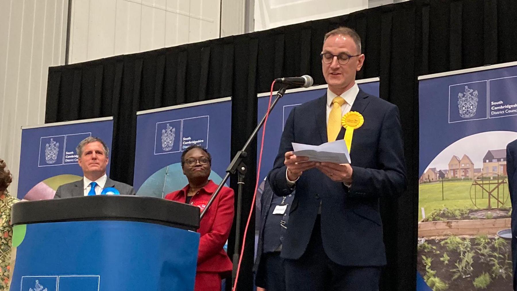 Ian Sollom giving his speech into a microphone behind a podium which reads "Elections", beside other candidates.