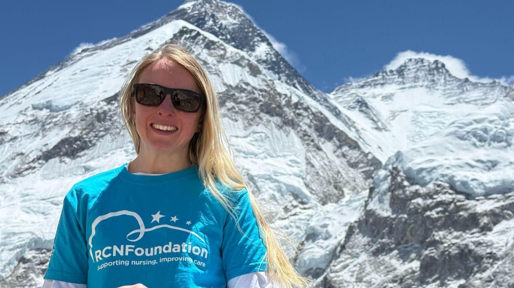 Rowena wearing a blue t-shirt with the RCN Foundation on the front. She is stood in front of a mountain wearing sunglasses. She has blonde hair.