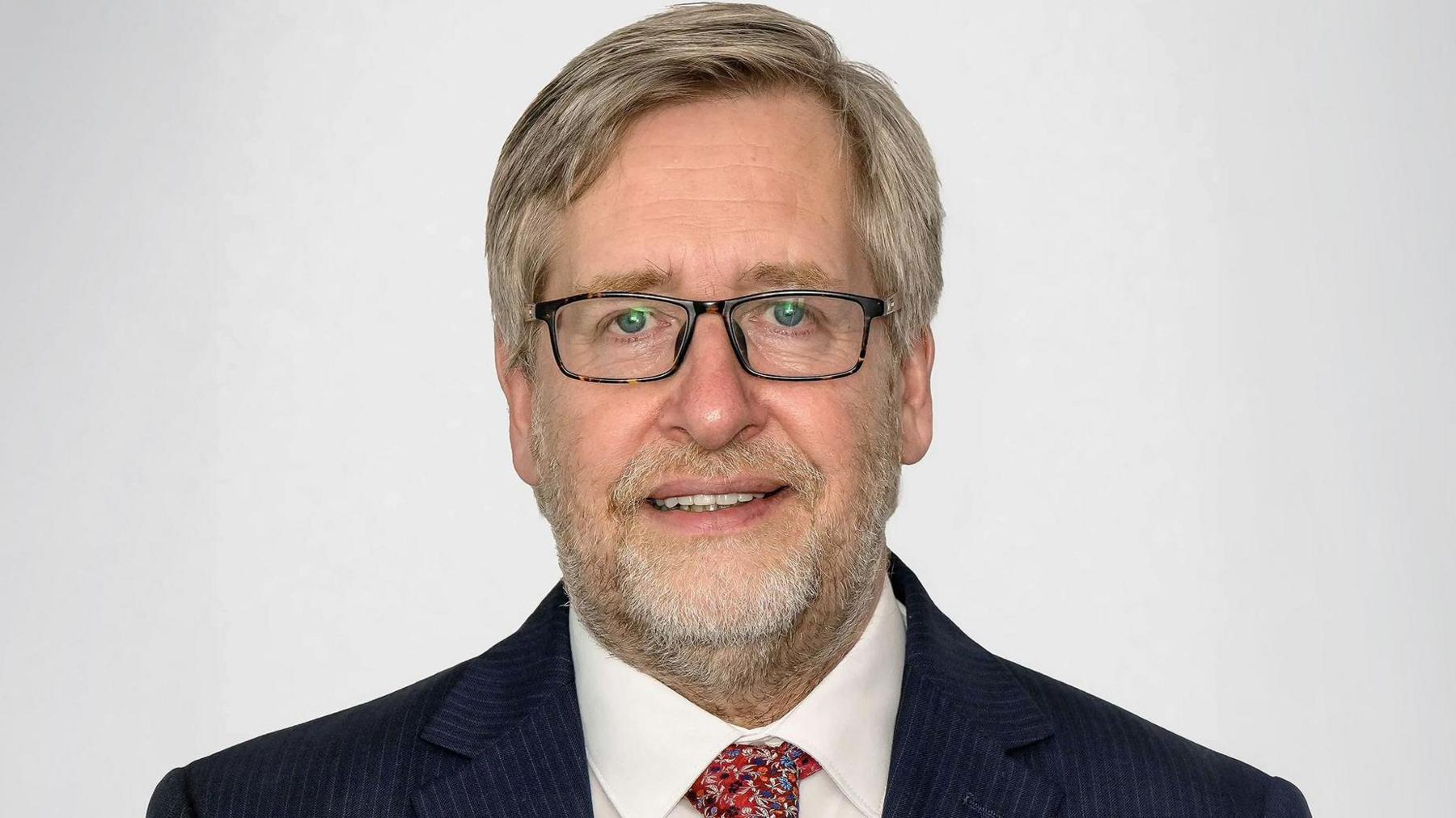 Man wearing a dark pinstripe suit with a white dress shirt and a patterned red tie, posed against a plain light gray background.