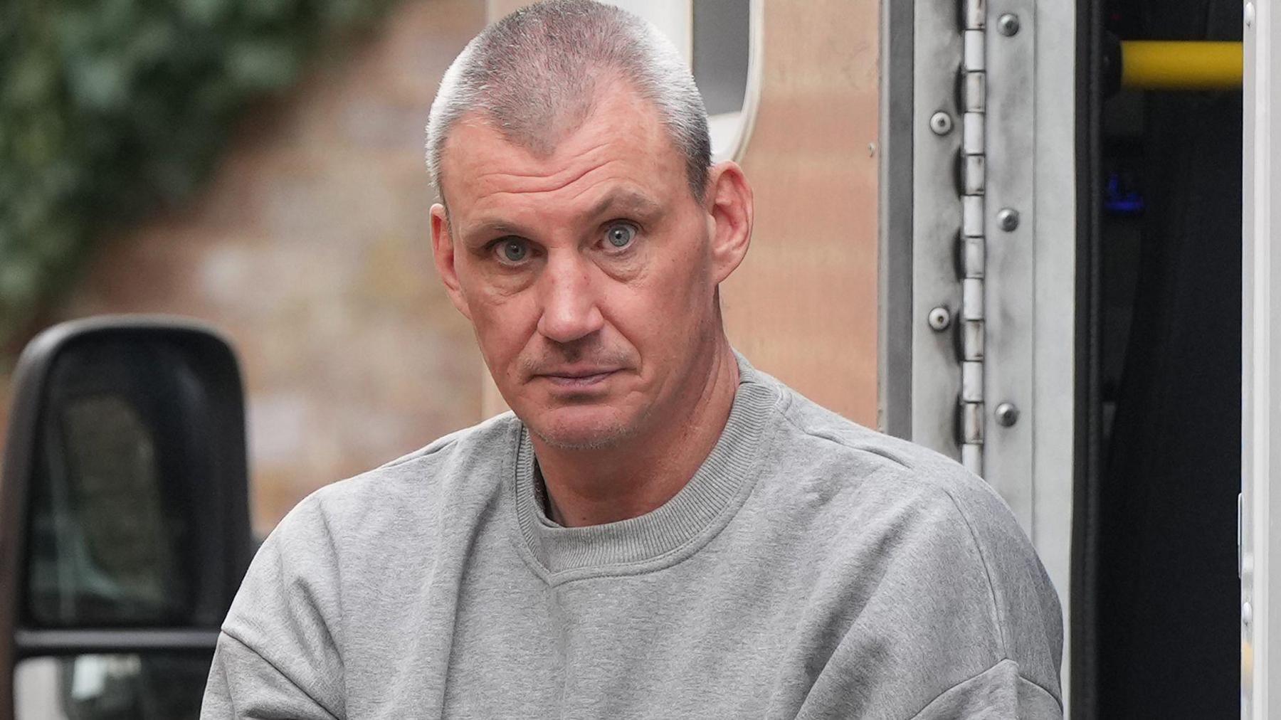 A man with short grey hair wearing a grey tracksuit top. He is being taken out of a prison van and staring at the camera.
