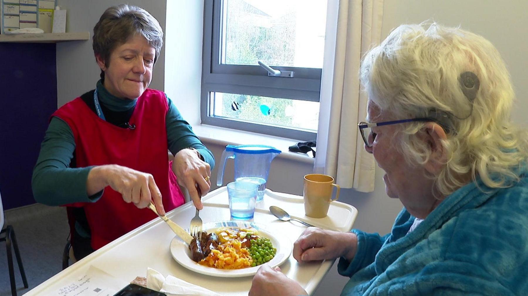 Volunteer Isabel helps a patient cut up some of her food in Newton Abbot Community Hospital. Isabel is wearing a red tabard. She is cutting some sausages for a woman in a blue dressing gown. 