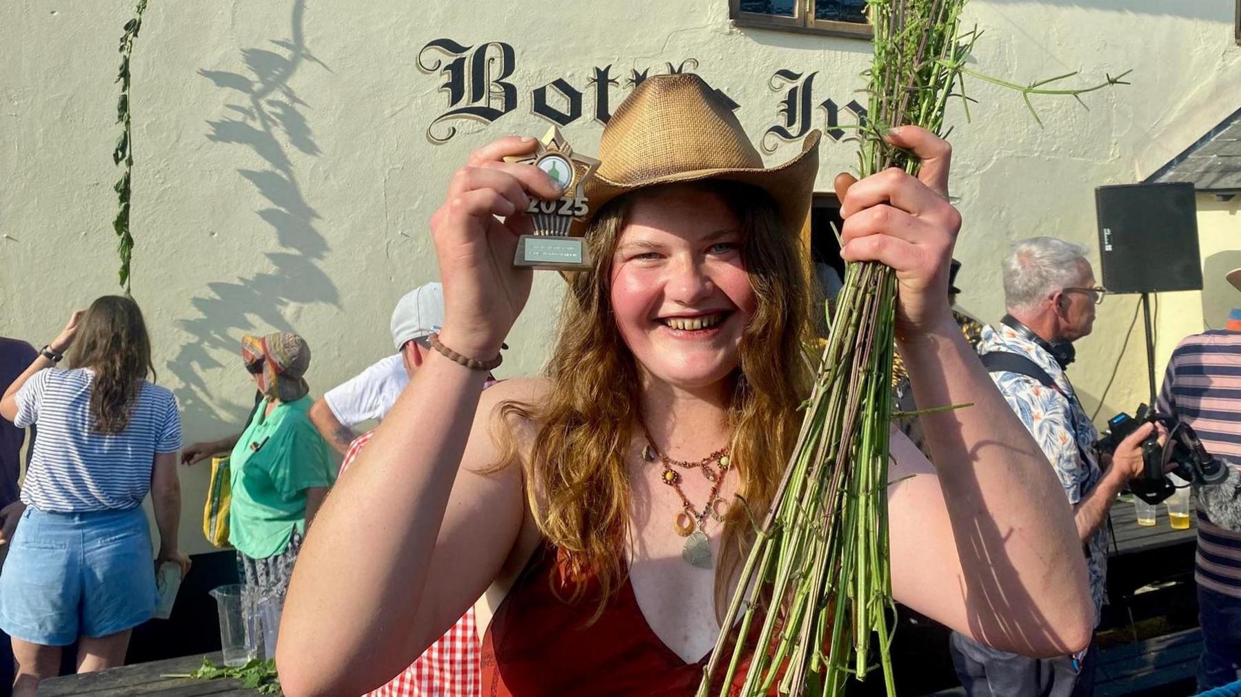 Lucy Dermody, wearing a straw hat, holding a bunch of plucked nettle stems, outside the thatched Bottle Inn pub