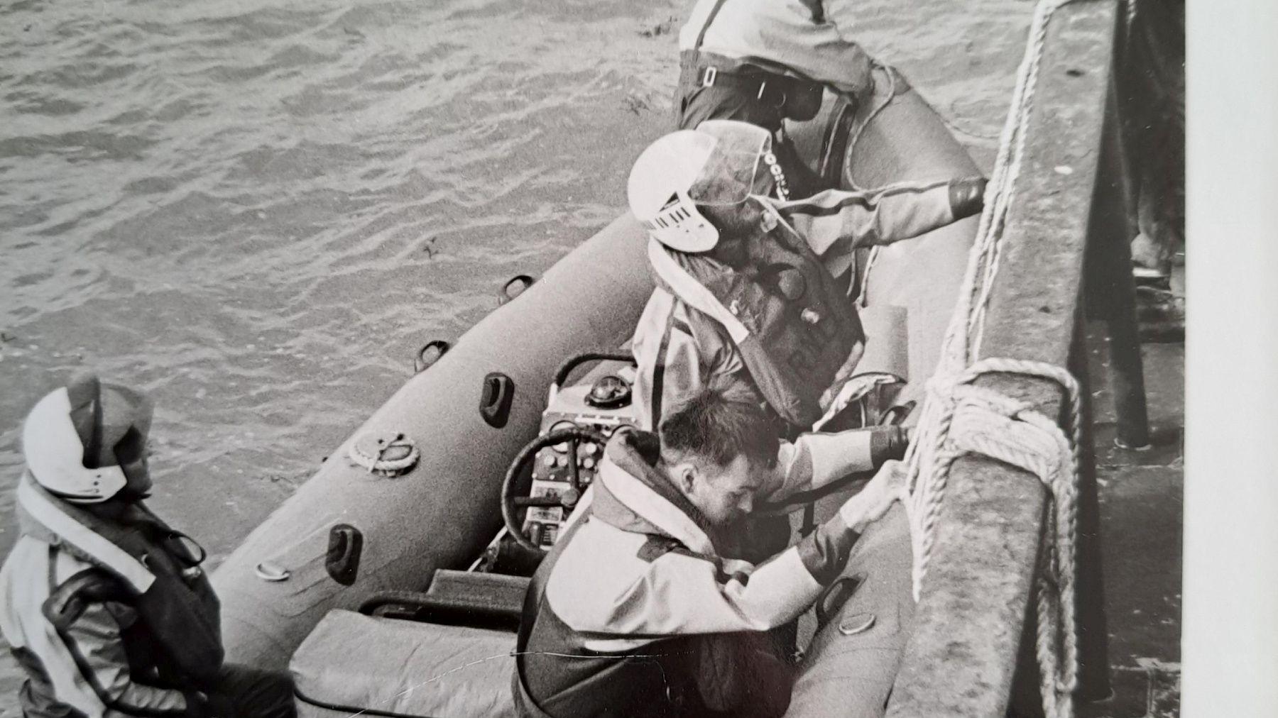 A black and white image of four coastguards on an RNLI lifeboat on water. There is some rope also in shot. 