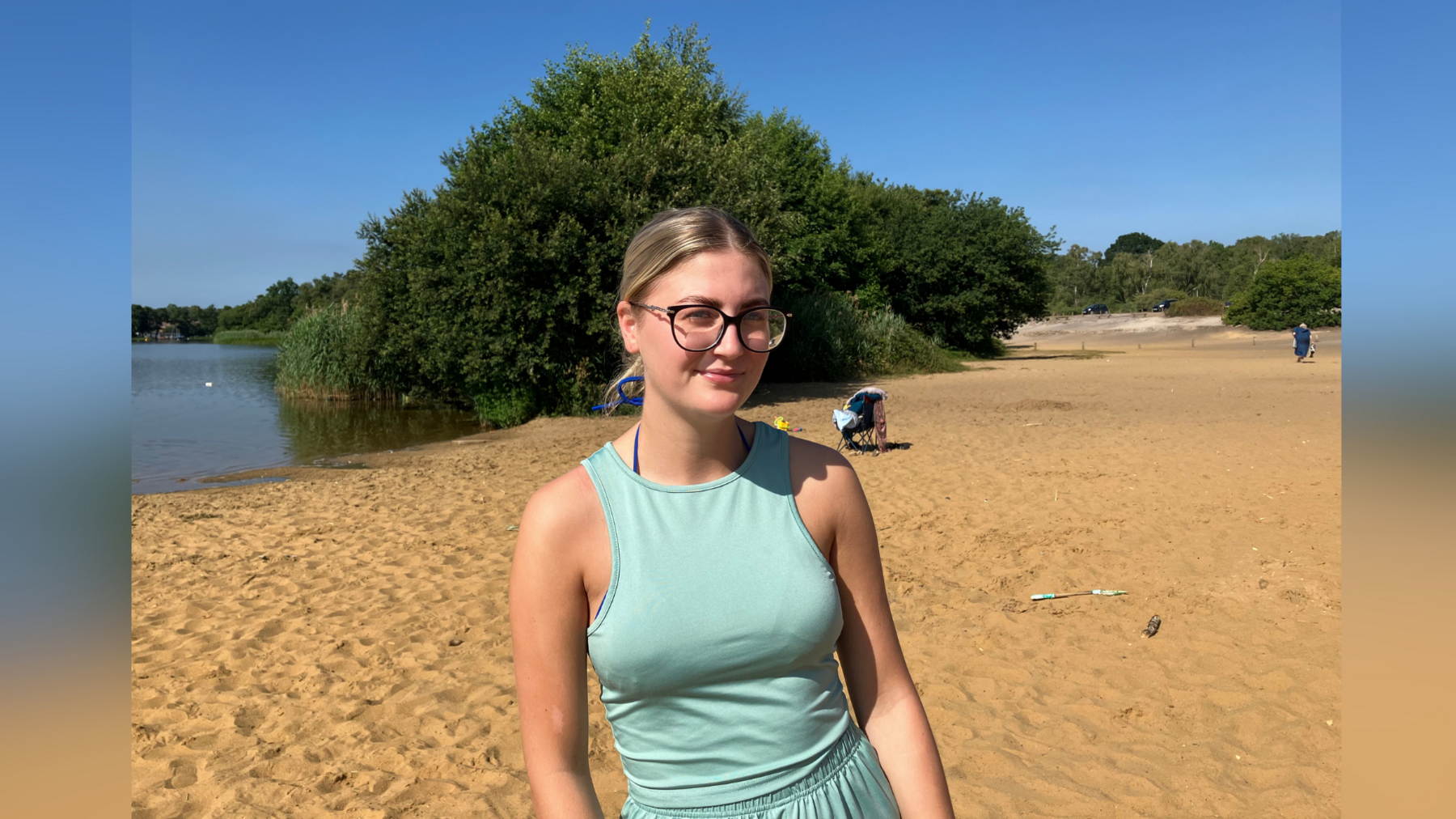 A picture of Tillie Dyer at Frensham Great Pond between Farnham and Hindhead, Surrey. Tillie is wearing glasses and a green top and shorts. Sand and lake can be seen in the background.