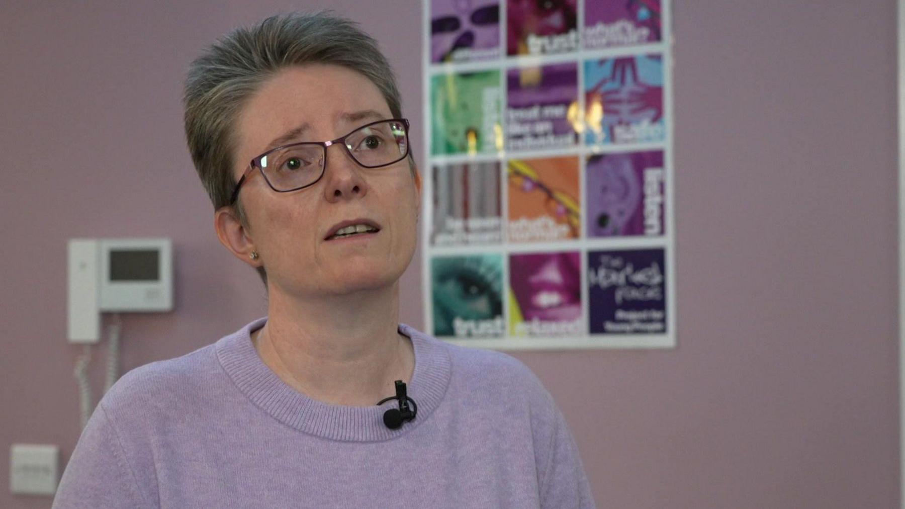 A woman with black-rimmed glasses and short grey hair. She is wearing a lilac jumper. A poster is on the wall behind her.