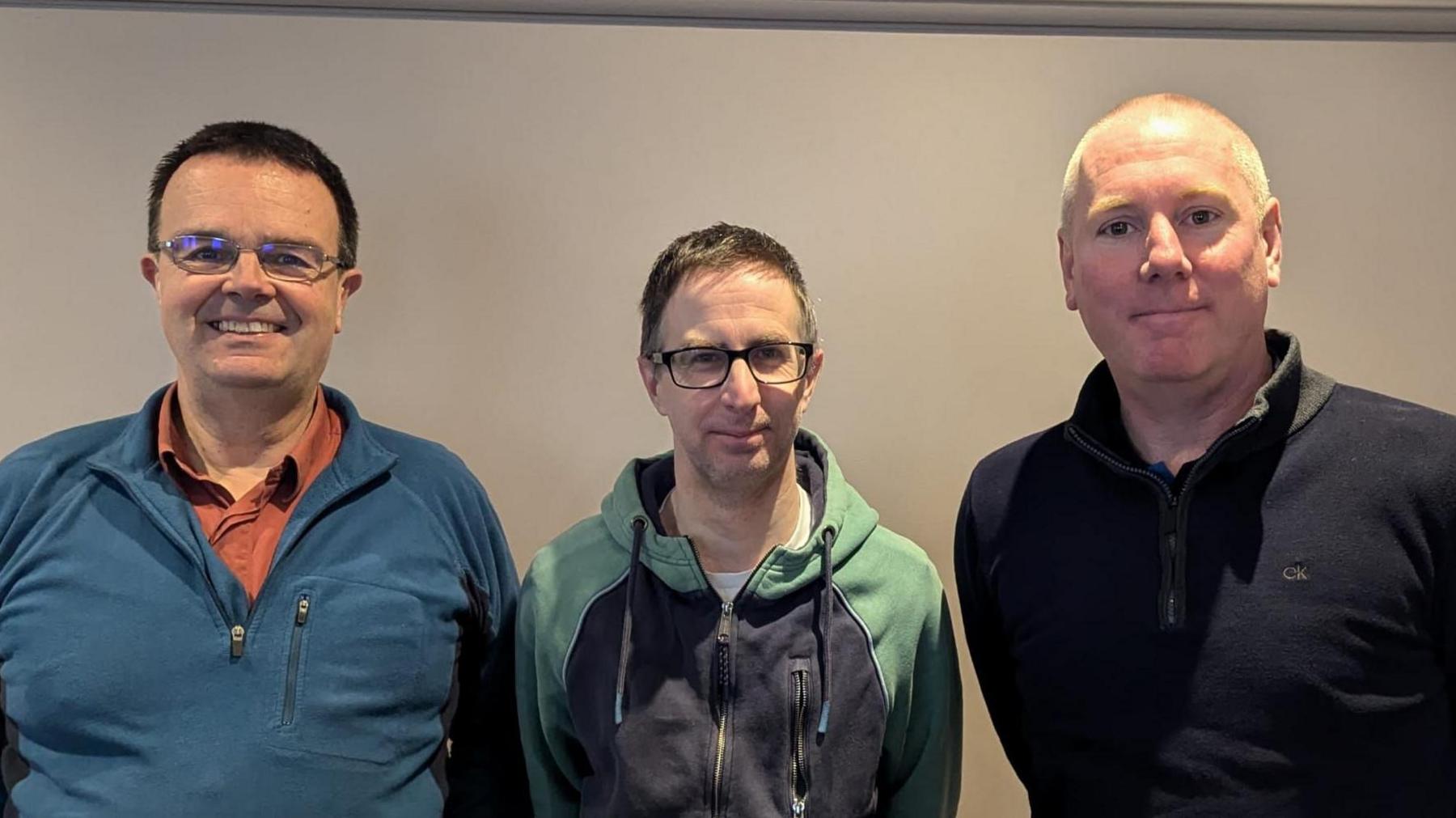 Three men look at the camera, the first is wearing a blue sweater and orange t-shirt, the second is wearing a blue and green sweater, and the third is wearing a dark top.