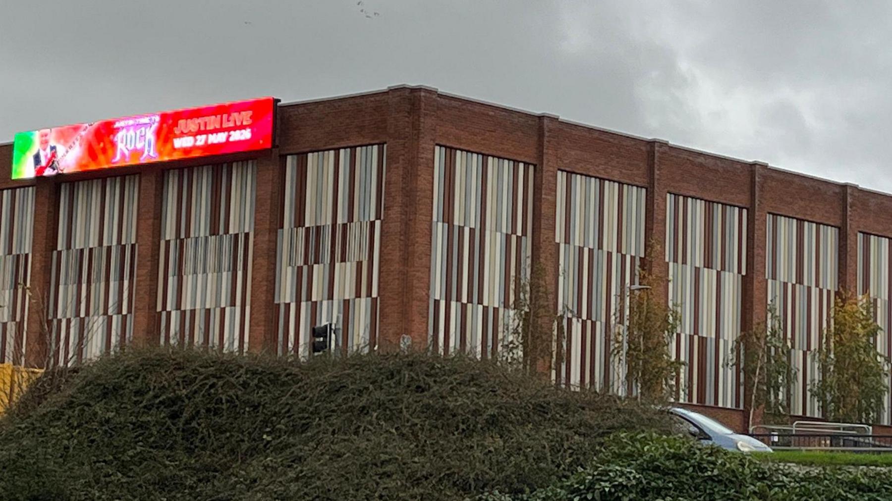 A general view of the Sands Centre in Carlisle taken from the pedestrian underpass opposite the building. The flat-roof building, pictured from the side where the leisure centre is, has a large LED screen on the front, advertising an event. A crane working on the side and back of the building is visible in the distance.