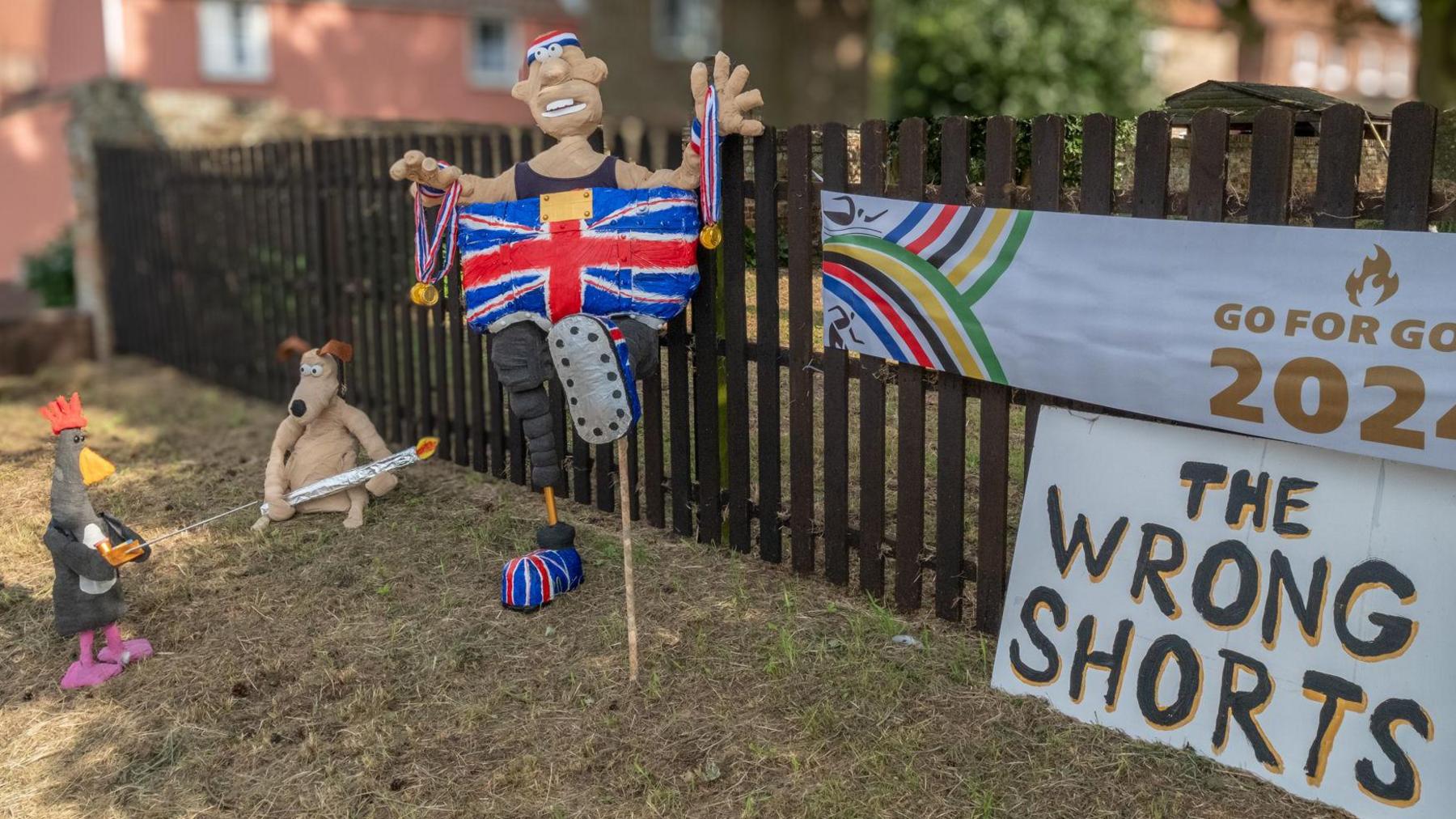 A papier-mâché of Wallace, Gromit and Feathers McGraw in front of a brown fence in a garden. Wallace is wearing Union Jack trousers as his arms raised while holding medals. Gromit is holding an Olympic torch and Feathers McGraw is holding a yellow remote control.