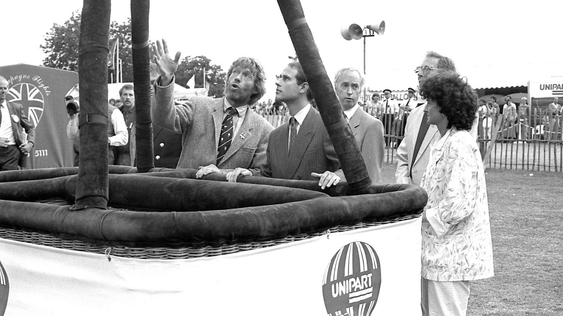 Black and white picture of Prince Edward looking up as a man standing next to him explains something to him. They are standing near the basket of a hot air balloon