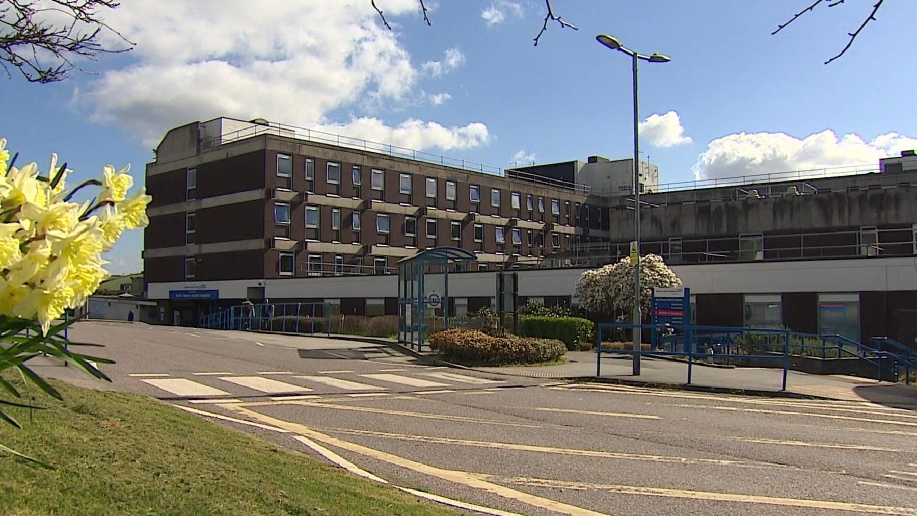 The outside of a hospital on a clear sunny day. The building has lots of windows and has the usual NHS branding on the front of it. There is a road in front of it with some flowers to the left of the pictures.
