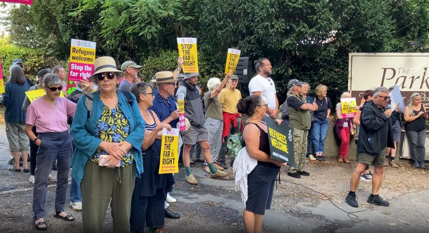 A group of about 20 people stand on a pavement area, holding yellow and pink placards saying Stop the Far Right and Refugees Welcome. Behind them is half of the sign for The Park Hotel.
