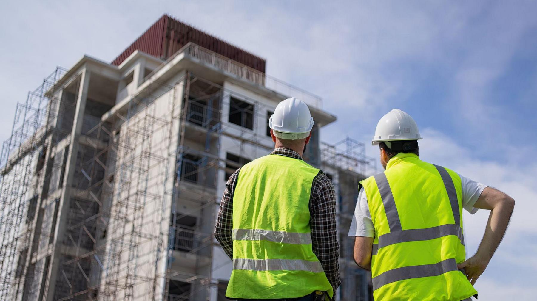Two builders in high visibility jackets and white hard hats look towards a tall set of unfinished flats with scaffolding around it.