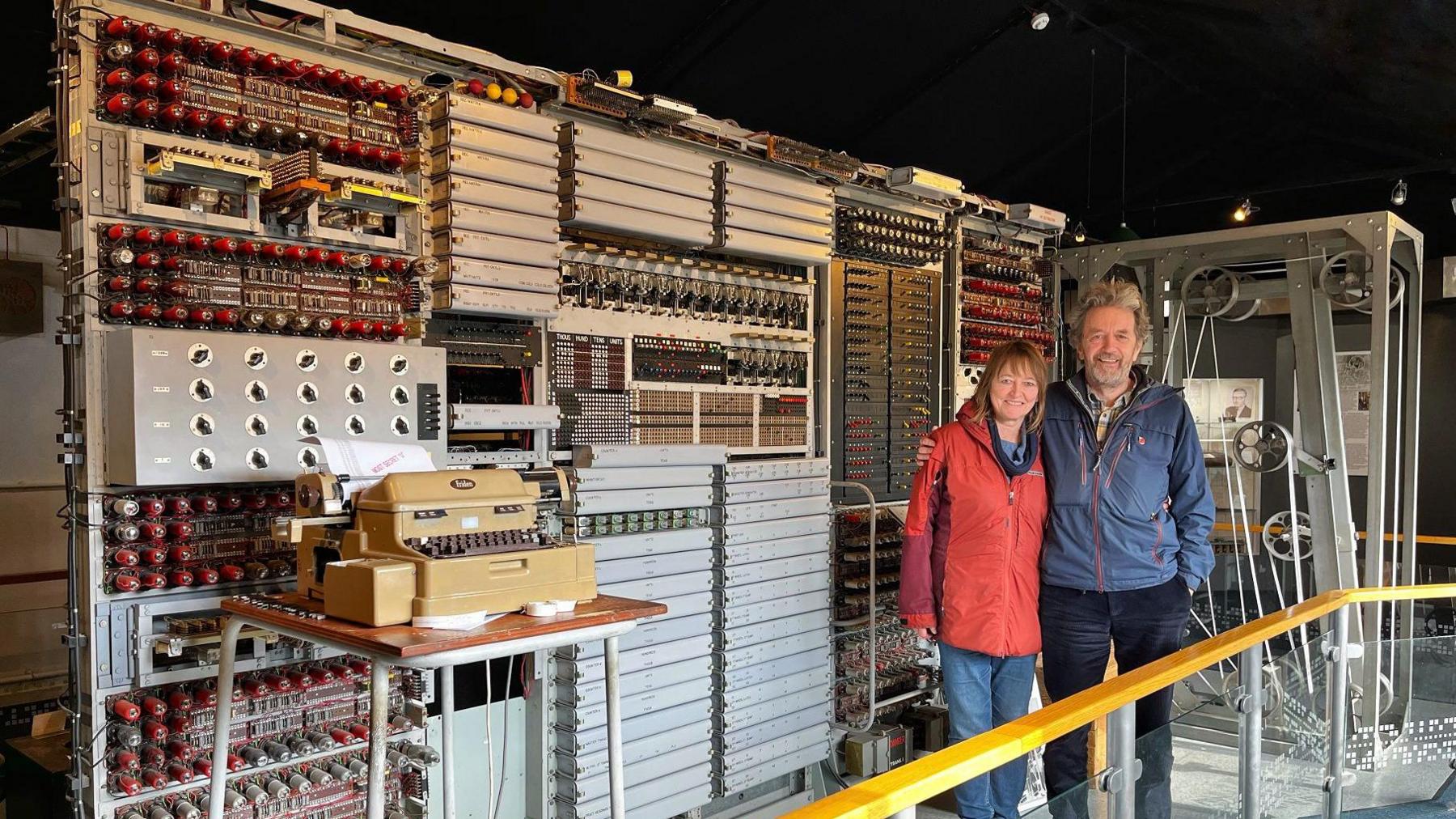 Peter Martin and his partner standing in front of the Colossus computer at the National Museum of Computing. Peter has grey tousled hair and a beard and is smiling cheerfully at the camera. He is wearing navy jeans and a blue coat. He has his arm around his partner, a woman with a bob haircut and fringe, wearing a red anorak and blue jeans.
The computer is a about 4m long x 1.5m tall and has multiple sets of valves and buttons. It is mostly silver with red valves.