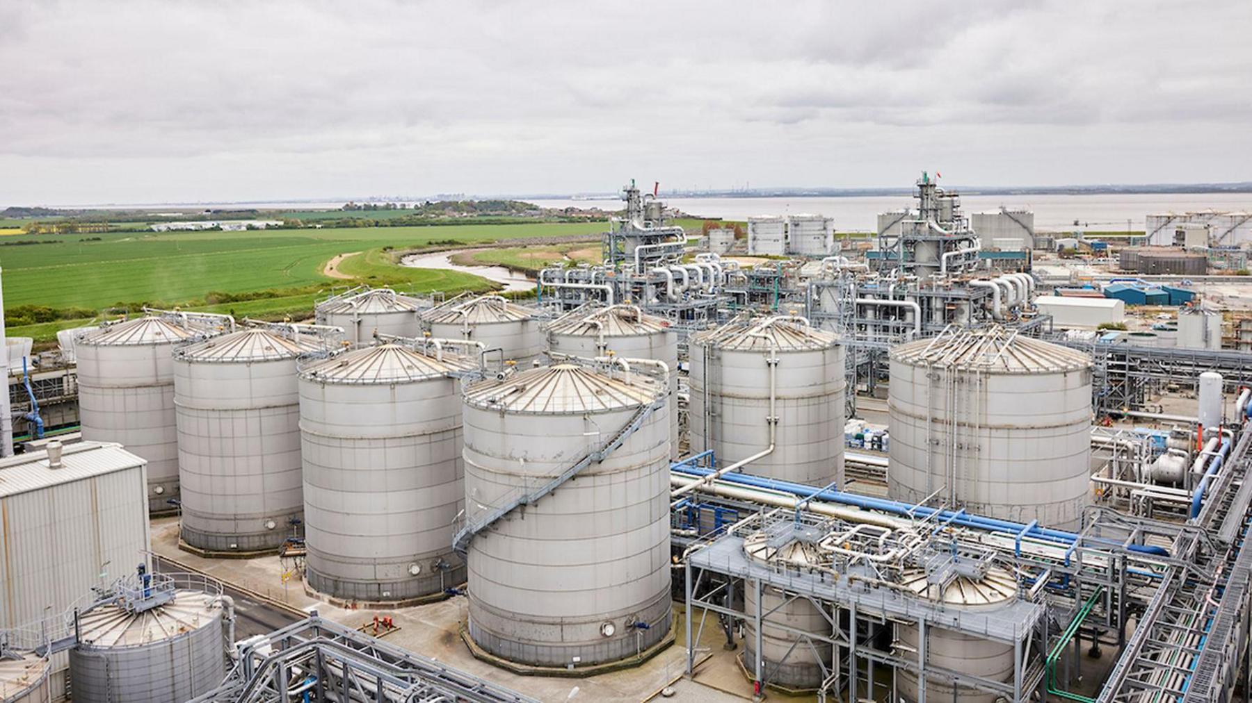 The exterior of the Vivergo Fuels plant, with huge metal cannisters at the front, metal railings and other grey metal buildings behind. A wide river estuary can be seen in the background, alongside fields and countryside.