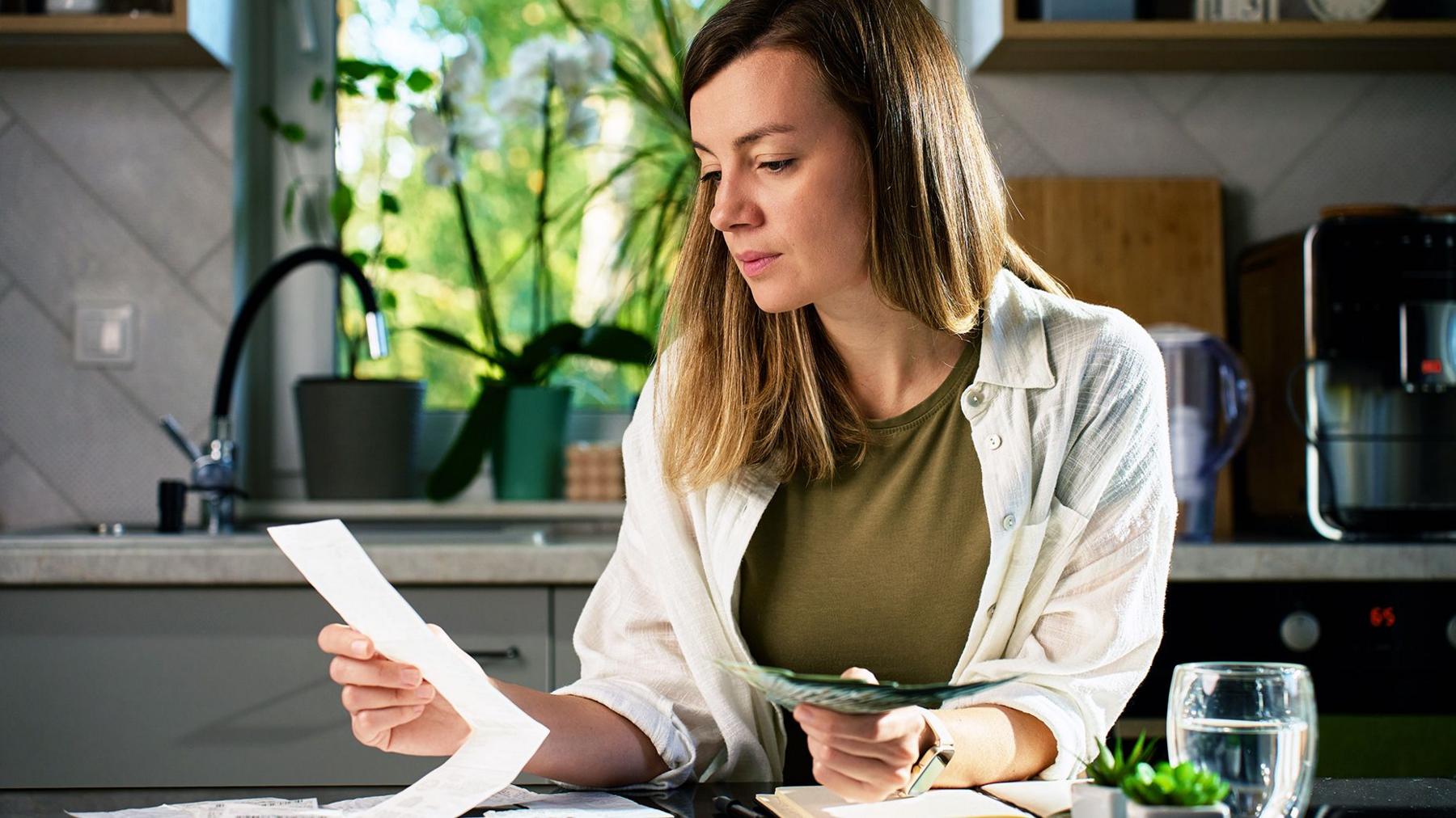 A woman sitting at a table and looking at houshold bills in her kitchen . In the background is a window showing green plants and trees