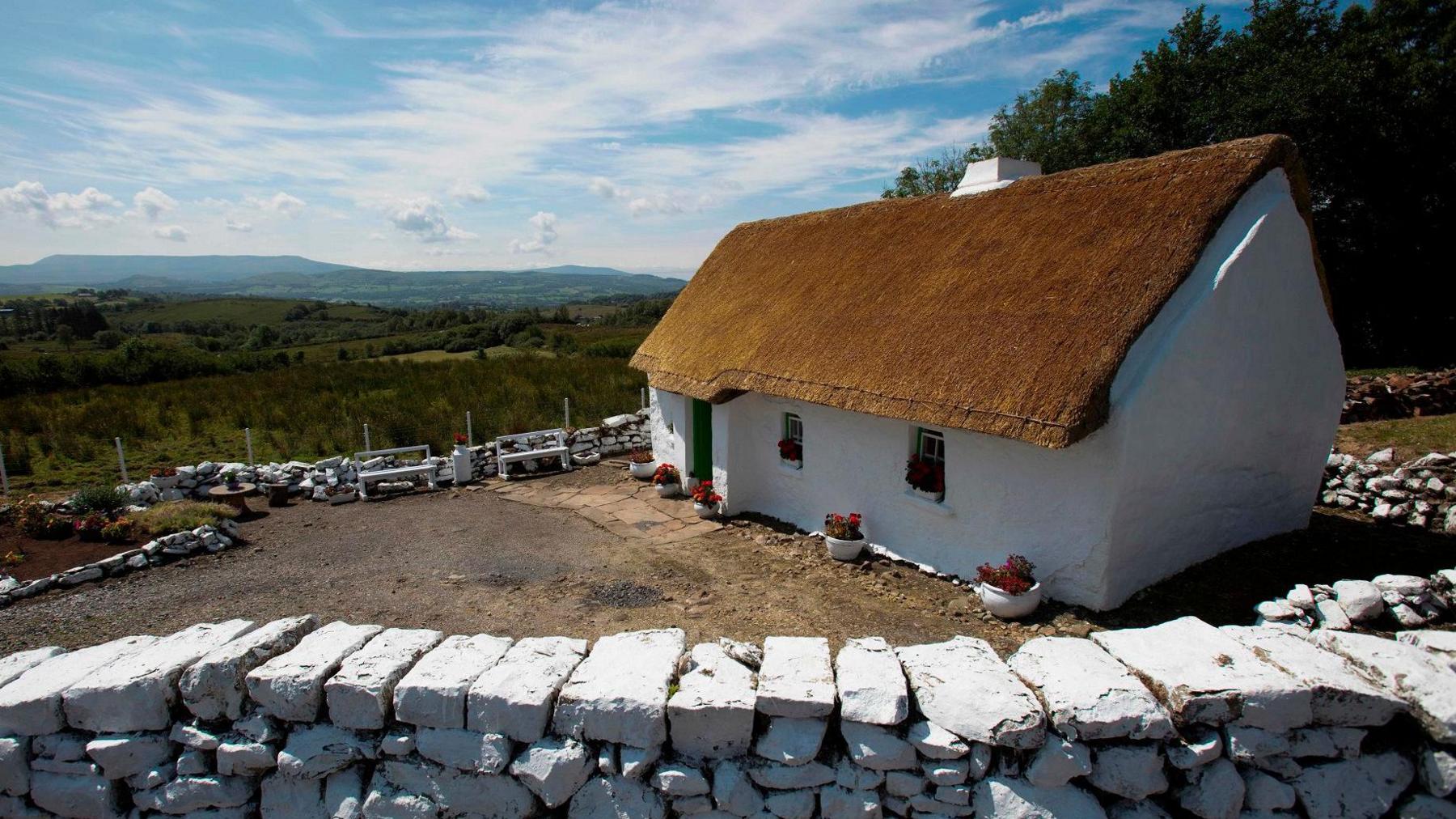 Mullylusty Cottage, a thatched home outside Belcoo in County Fermanagh.  The small three-room building is white washed and has three windows and a door at the front. There is a dry stone wall around the house, which overlooks fields and distant hills. 