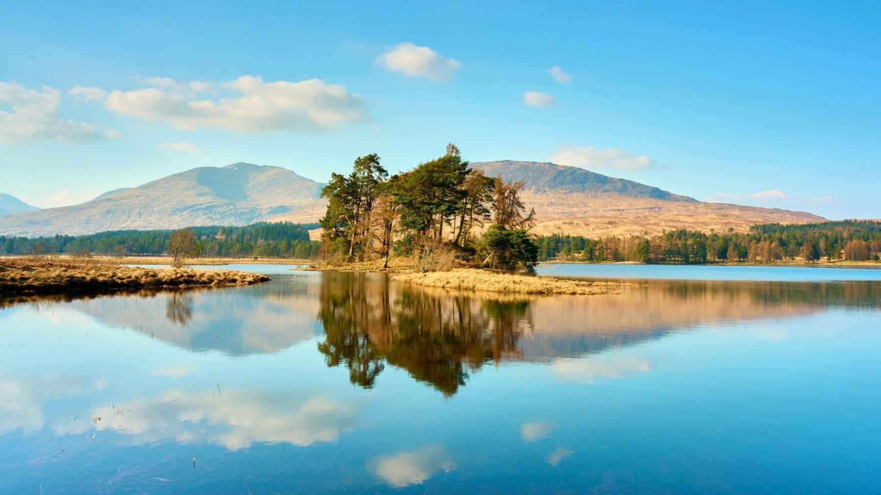 A general view of Loch Tulla. The sky is blue with a few clouds. It is reflected in the water. There is a small island in the middle with a few trees. There are mountains in the background.