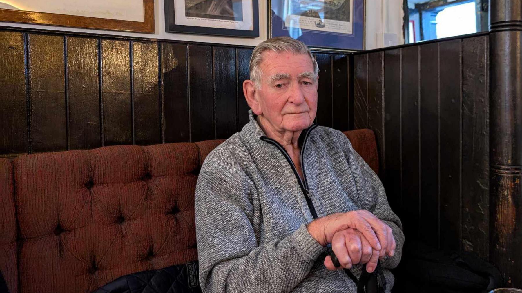 Tom Webb, who is 89, sitting in the Low Lights pub, wearing a grey knitted, zip up hoodie and holding both hands in front of him on the top of a walking stick.