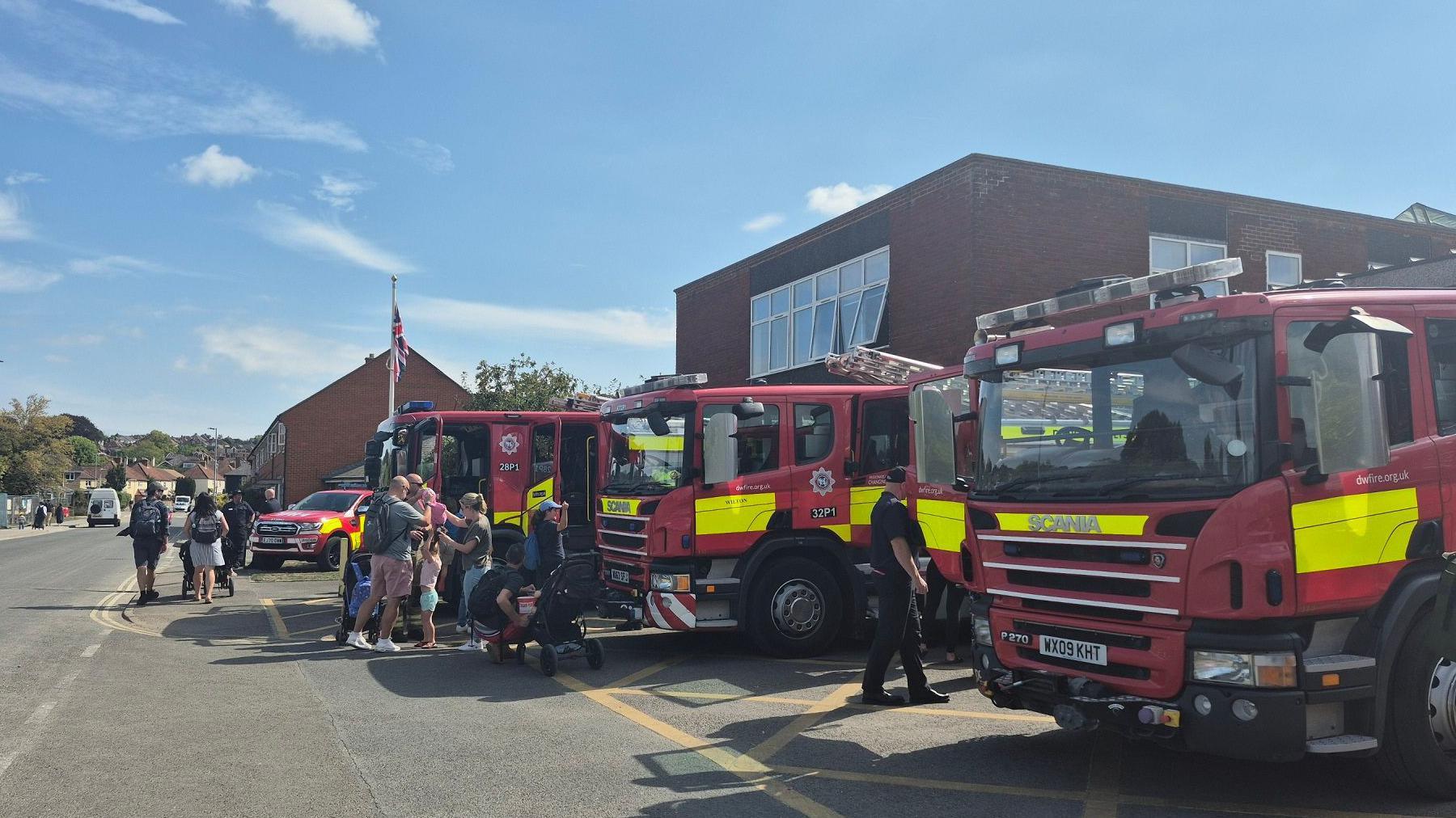 A row of fire engines parked outside a fire station along a main road. A gathering of people is on front of one. A sunny day with blue sky.