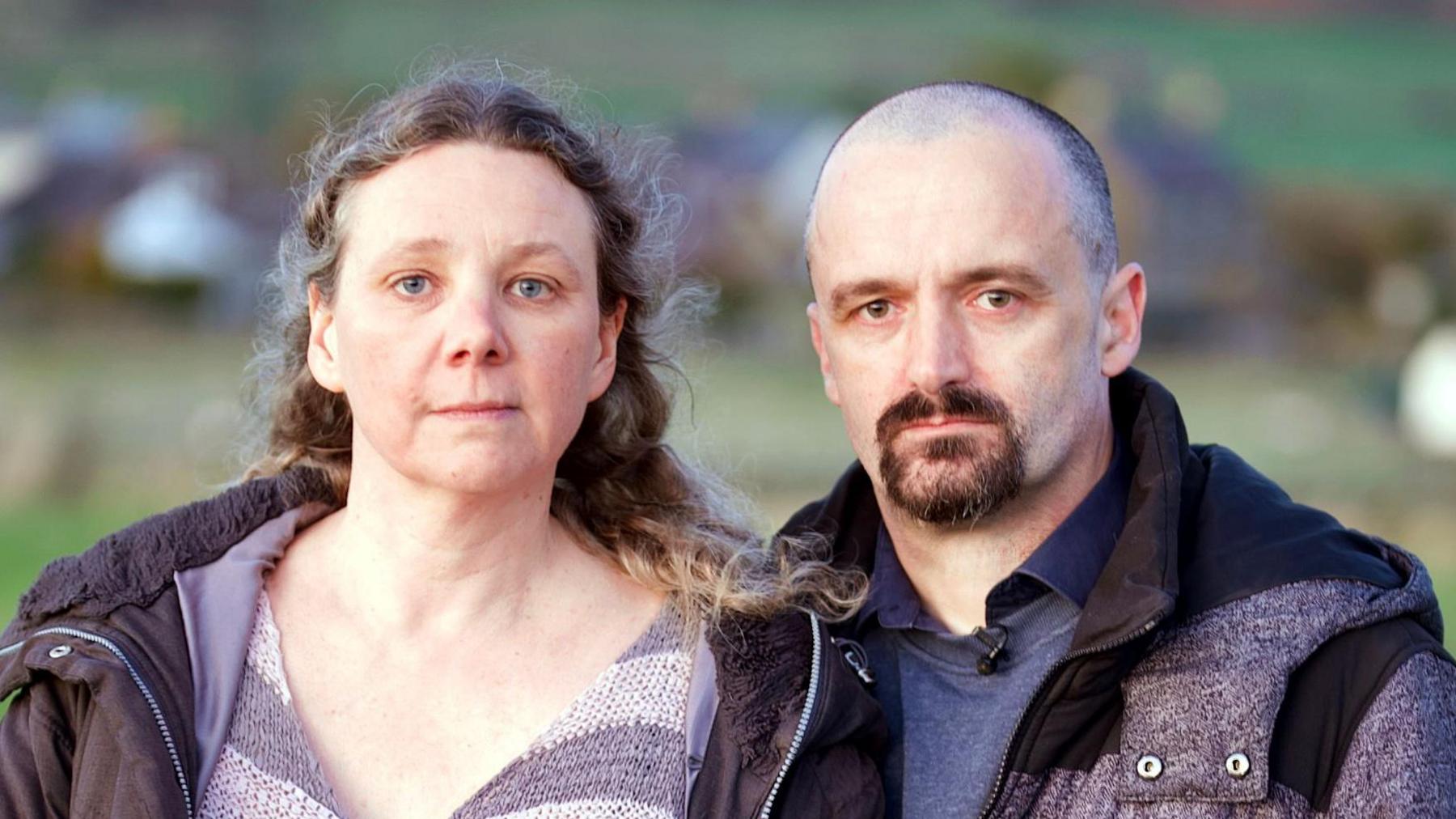 Close-up portrait of a couple standing outdoors in the countryside, with houses and a hillside in the blurred background behind them. Verity is a woman with long, brown, curly hair, blowing a little in the wind, who wears a striped jumper and a black coat with the hood down. Her husband Ian has a shaved head and a brown goatee beard, and wears a grey coat.