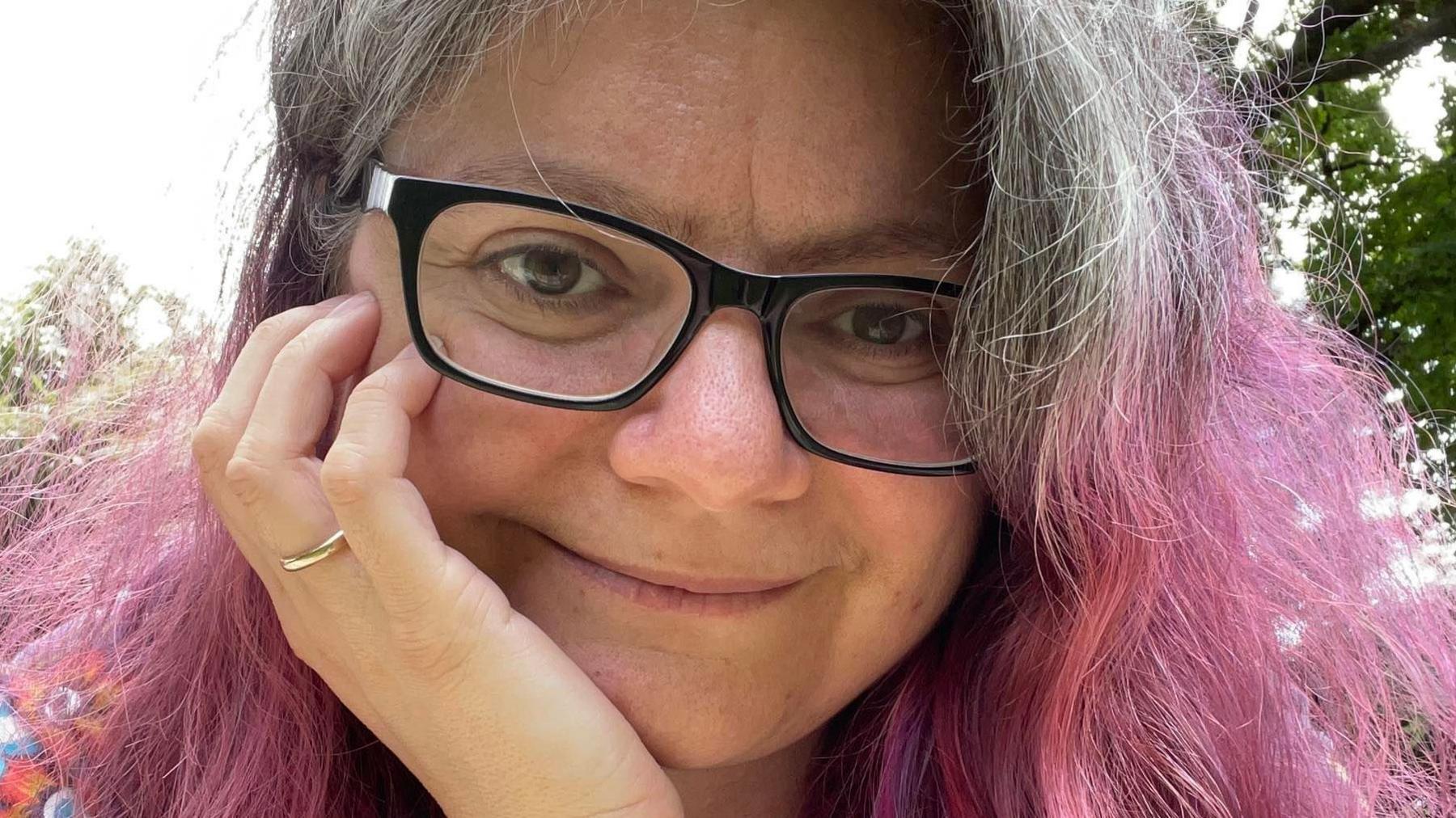 Dimita Fimi, who has wavy grey and dyed-pink hair and is wearing glasses, smiling and looking at the camera with her chin propped on her hand