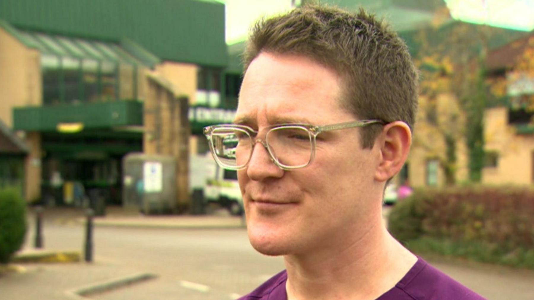 Dr Charlie Beattie wearing glasses and a purple top.  He is being interviewed outside Antrim Area Hospital