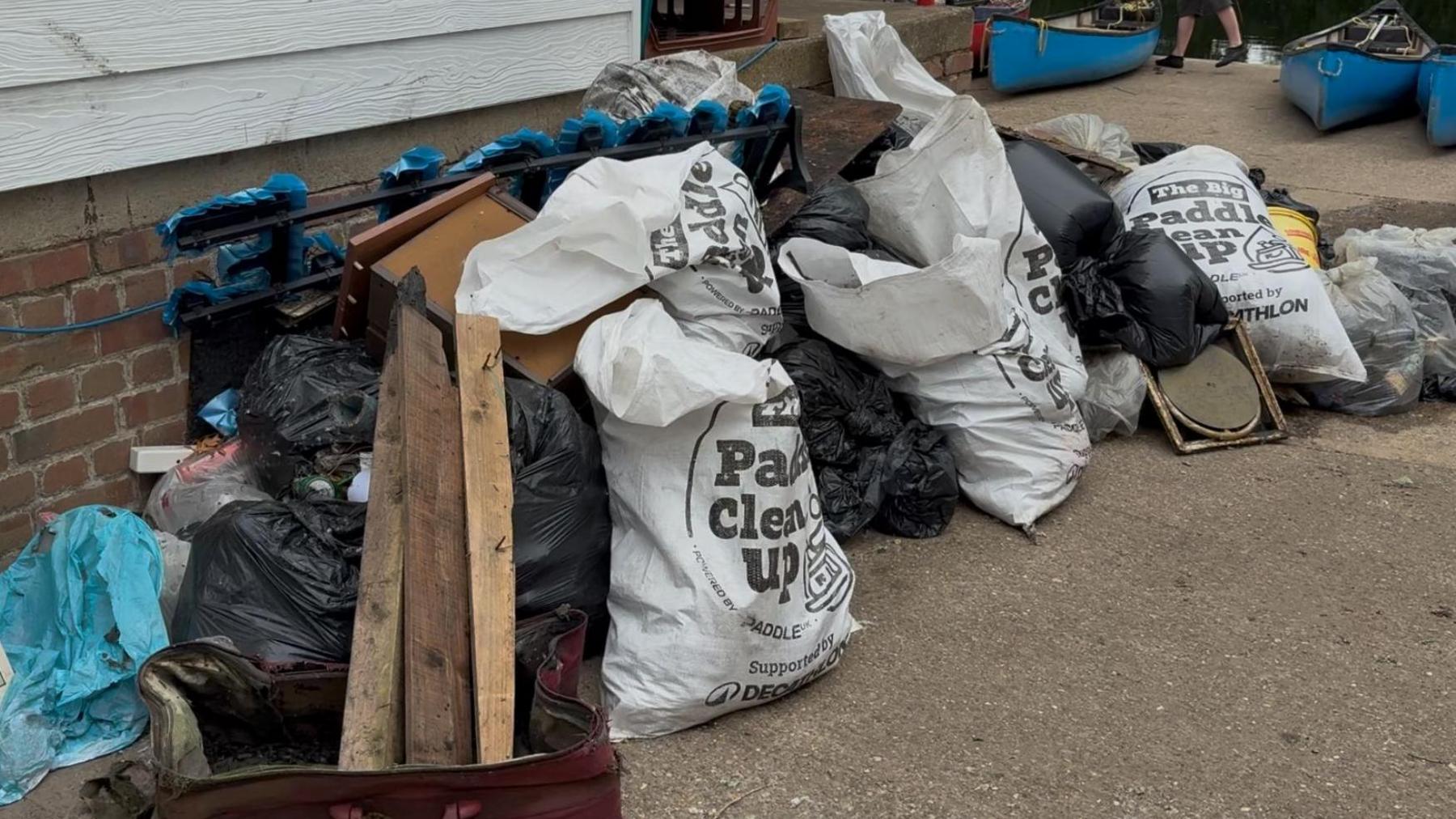 A pile of rubbish in bags sits at the side of a river next to three canoes.