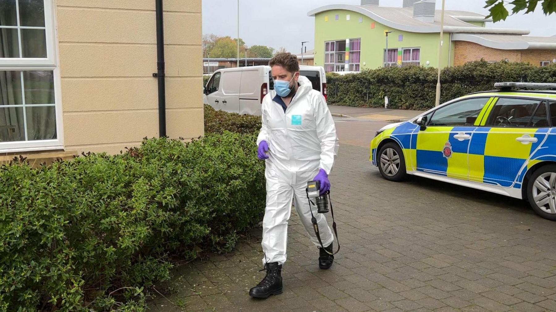 A man wearing a white forensic suit, a blue face mask, purple latex gloves and black boots. He is holding a camera in his left hand and walking towards a flat which has a large bush below the window. Behind him there is a parked police car with hi-vis yellow and blue decals. 