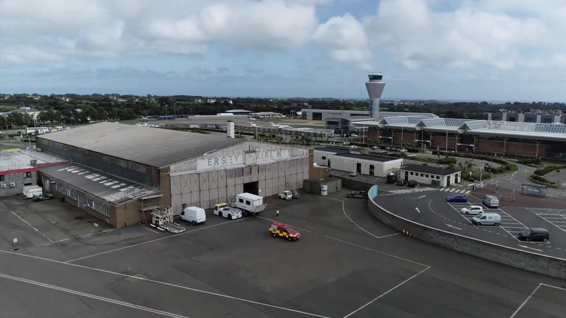 Large grey hangar building with old sign where some of the letters are no longer visible. Air control tower and other airport buildings visible in the background