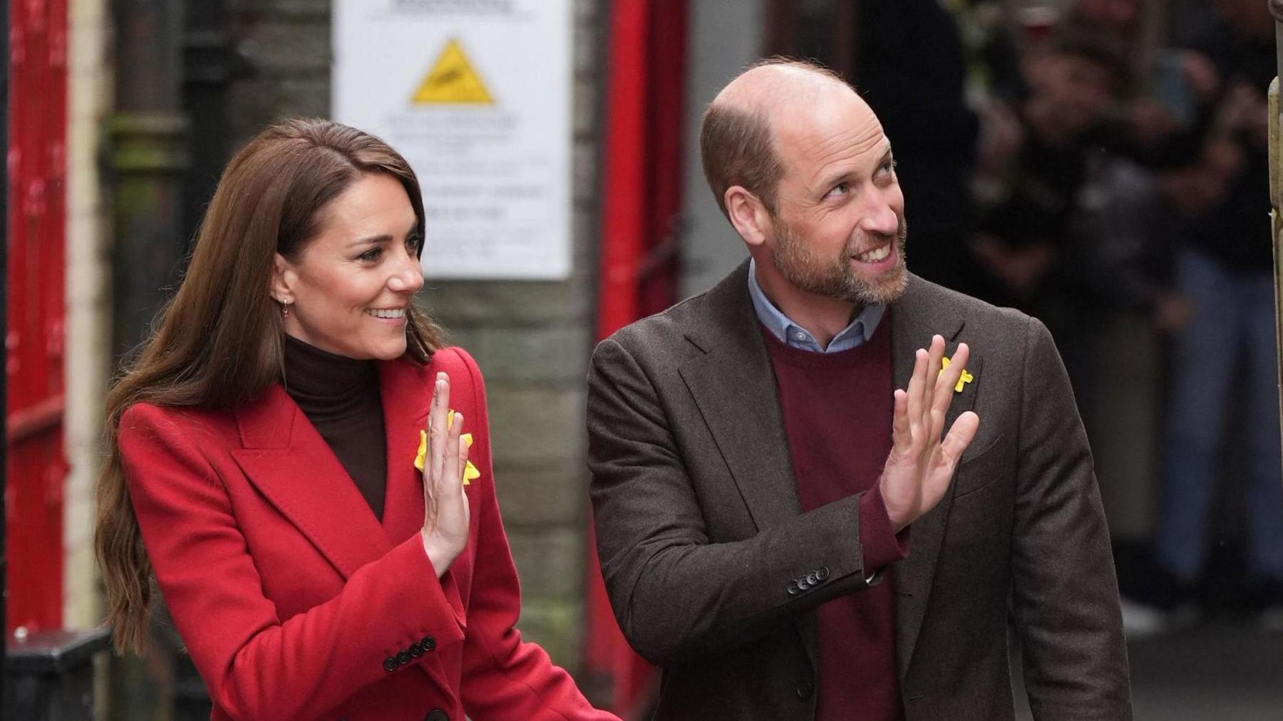 The Prince and Princess of Wales at Pontypridd Market walking through the town. Both are waving at crowds waiting on the right out of view of the camera. Catherine is wearing a red coat with four black buttons on the sleeve, she has long brown hair and is wearing a large daffodil pin on her lapel. William is wearing a blue shirt with a maroon jumper and a brown suit jacket. He is also wearing a daffodil pin on his lapel. 